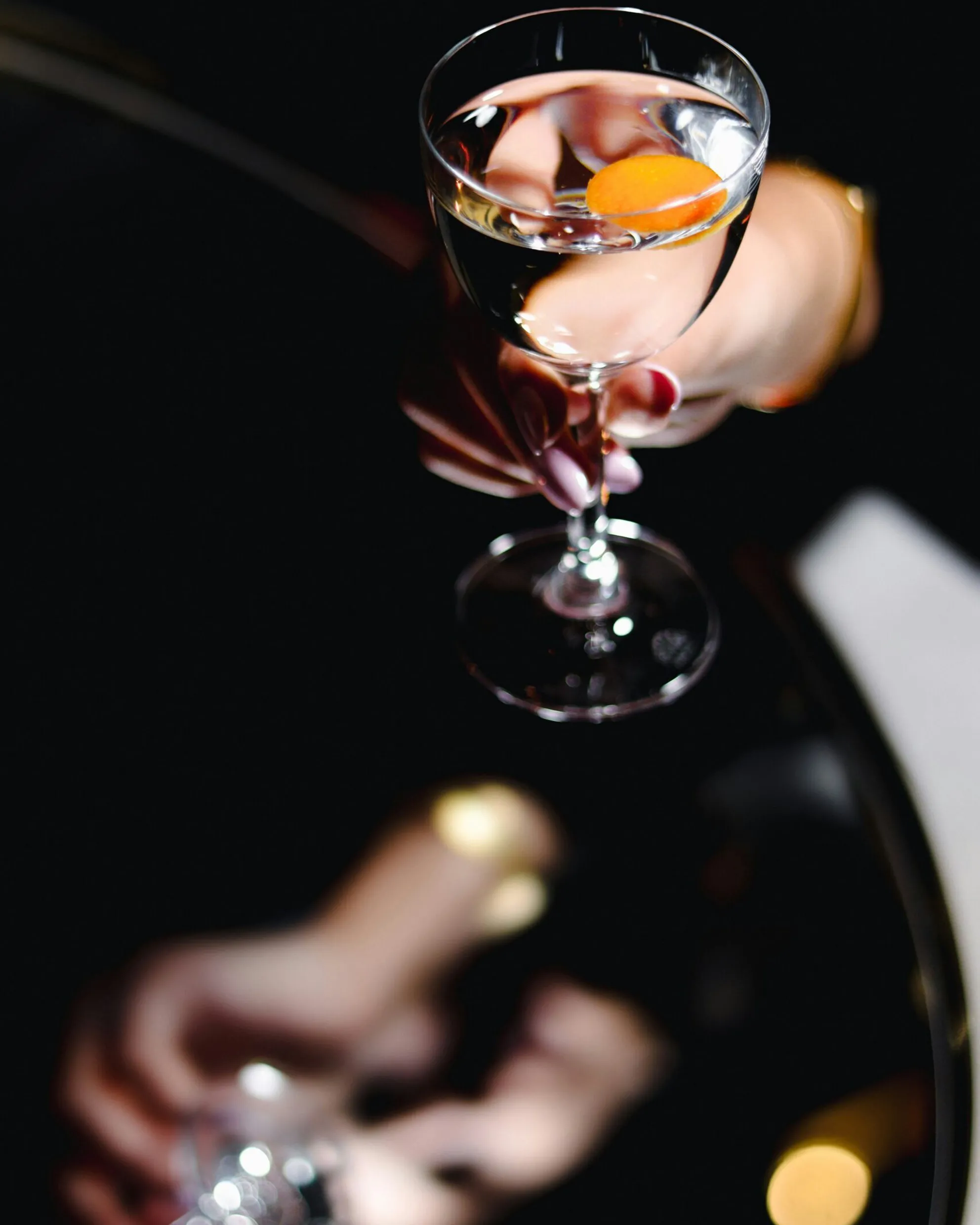 Martini glass with an orange peel, held in the hand of a woman with red nail polish, soft lighting.