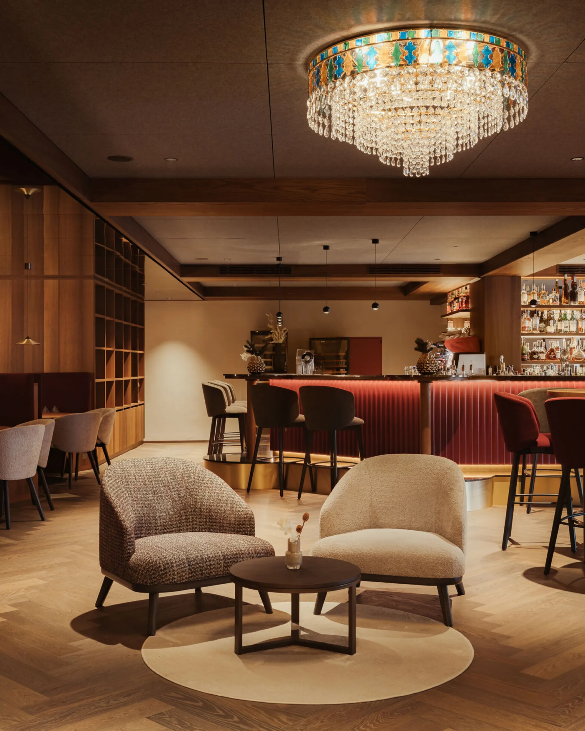 Warmly lit hotel bar with plush seating, a round table, and a chandelier. A wooden bar with red chairs creates a cozy, inviting atmosphere.