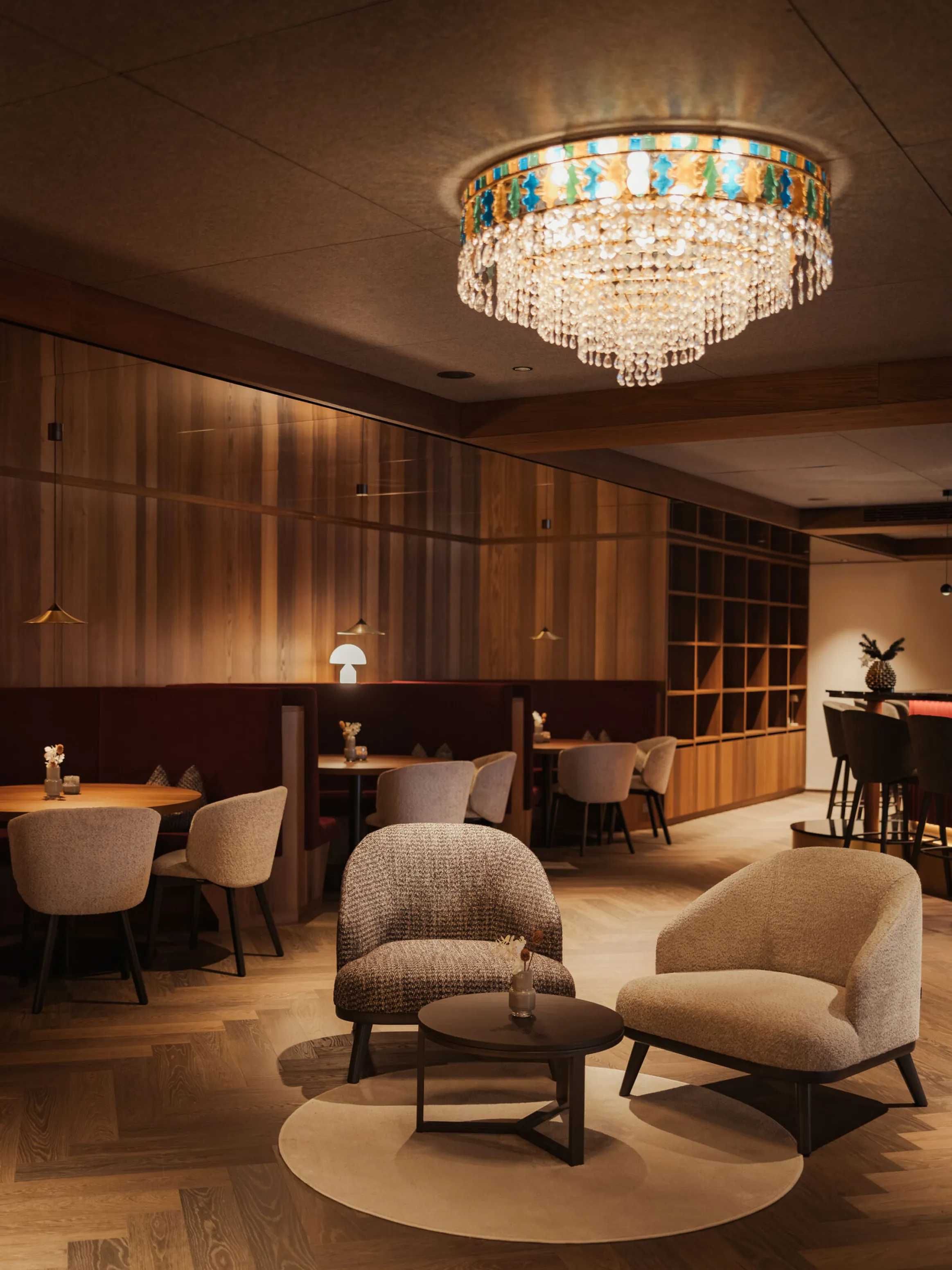 Cozy, ambient cafe interior with soft lighting. Features plush chairs, small round tables, a wooden wall, and an elegant crystal chandelier overhead.