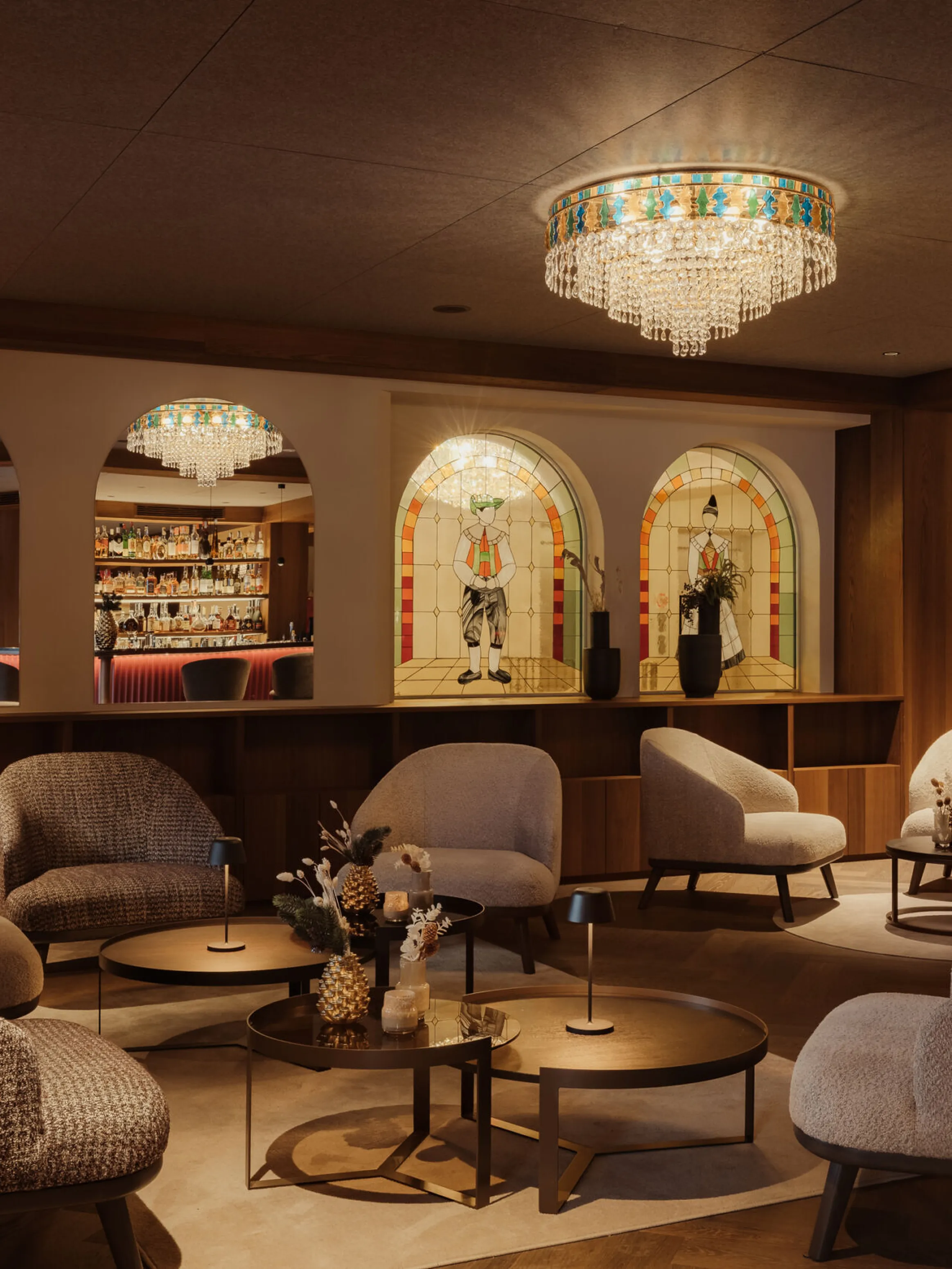 Cozy lounge with soft gray chairs around round tables, under a sparkling chandelier. Arched mirrors and a lit bar create a warm, inviting atmosphere.