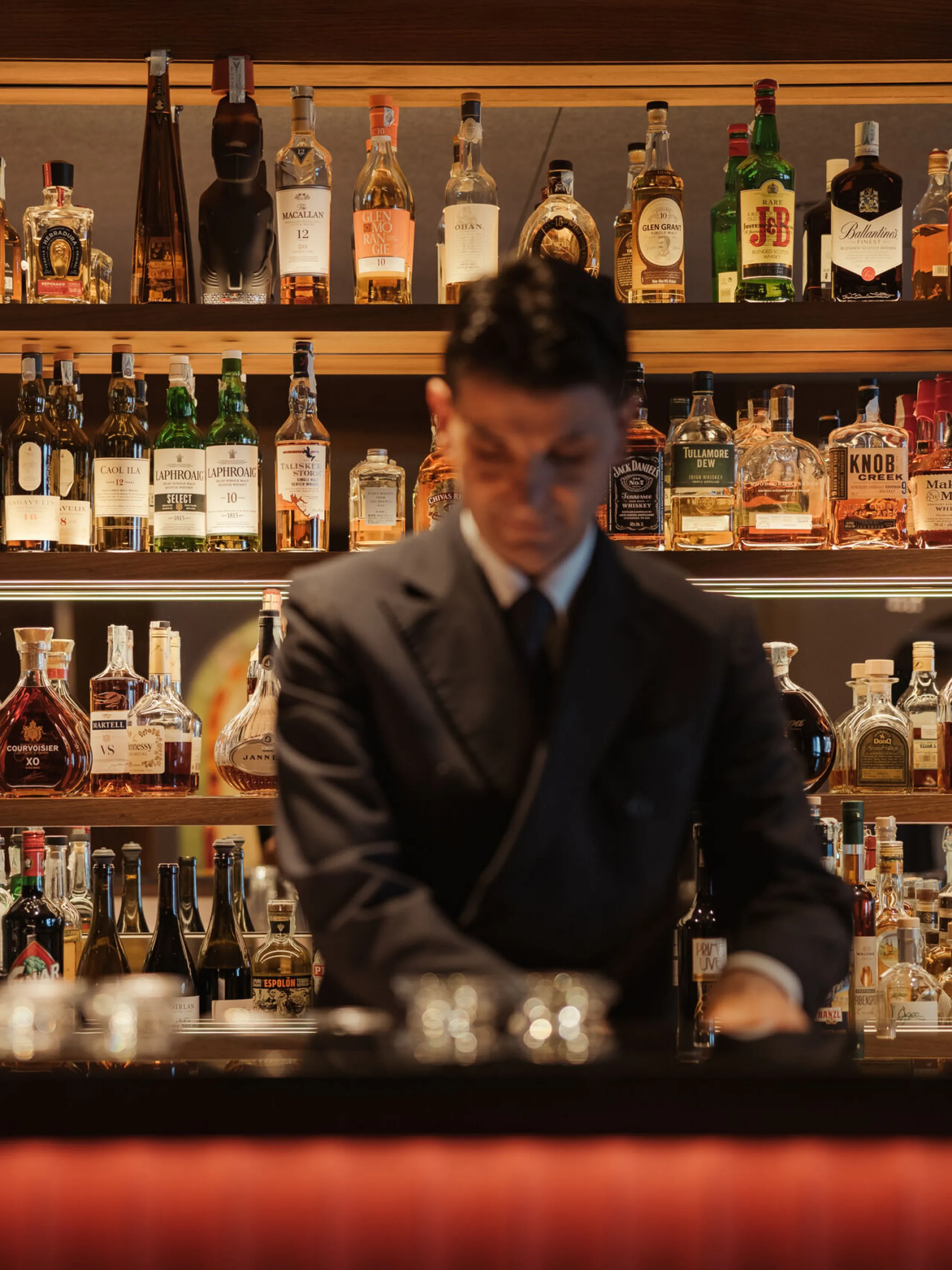 A bartender in a suit stands at a dimly lit bar, surrounded by shelves stocked with an array of liquor bottles, creating an elegant and sophisticated ambiance.