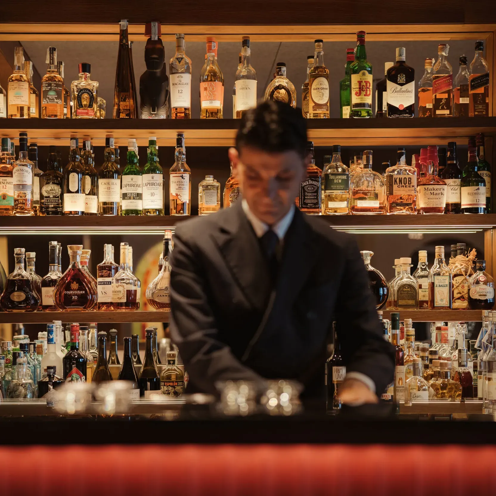 A bartender in a suit stands at a dimly lit bar, surrounded by shelves stocked with an array of liquor bottles, creating an elegant and sophisticated ambiance.
