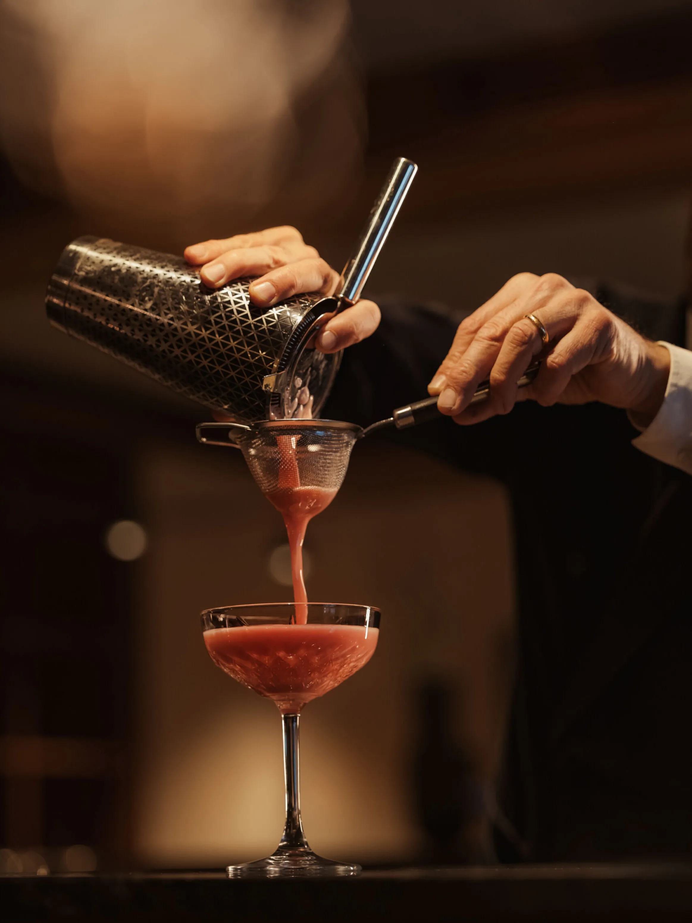 A bartender pours a vibrant pink cocktail into a glass, using a shaker and strainer. The warm lighting creates an elegant, sophisticated atmosphere.