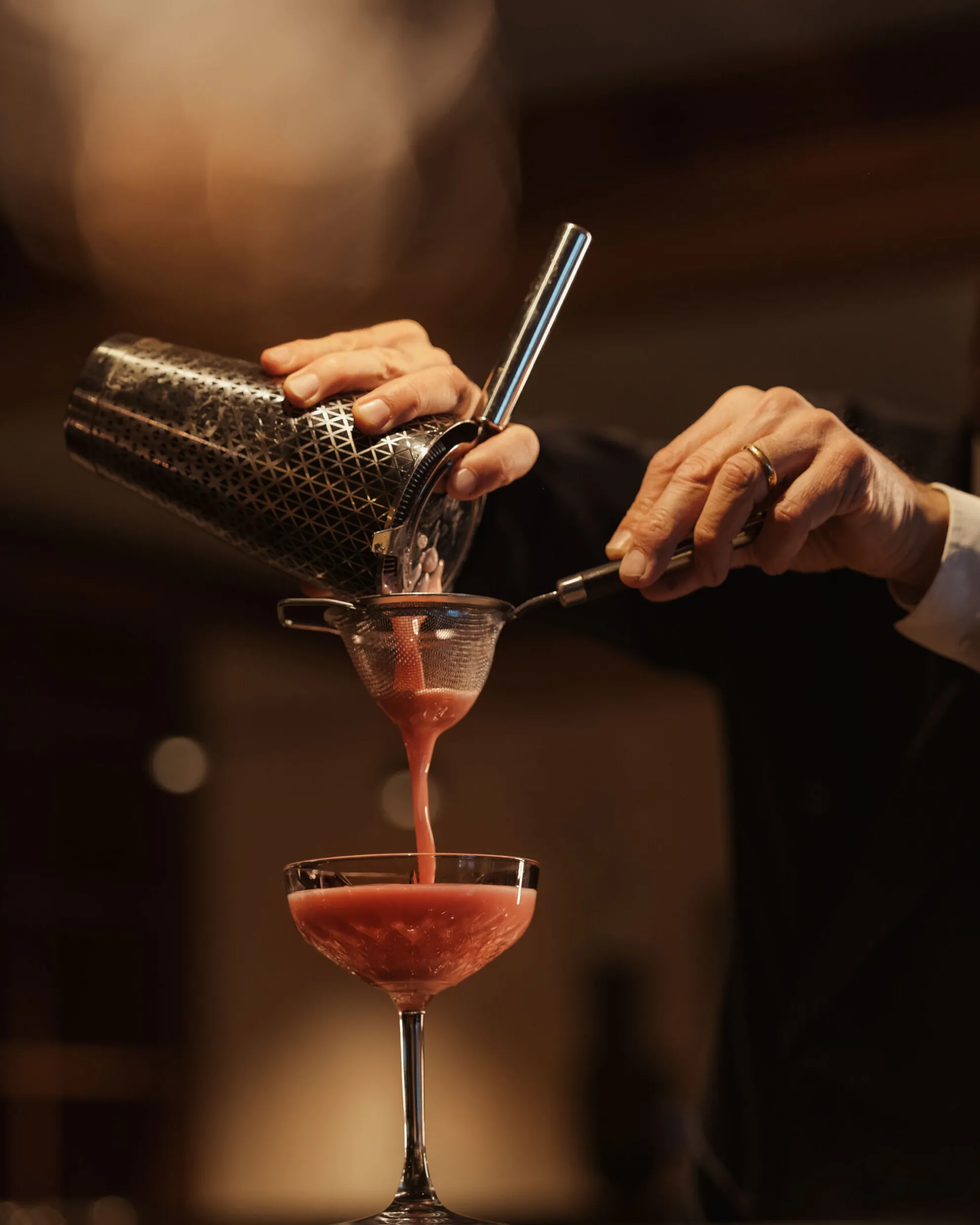 A bartender pours a vibrant pink cocktail into a glass, using a shaker and strainer. The warm lighting creates an elegant, sophisticated atmosphere.