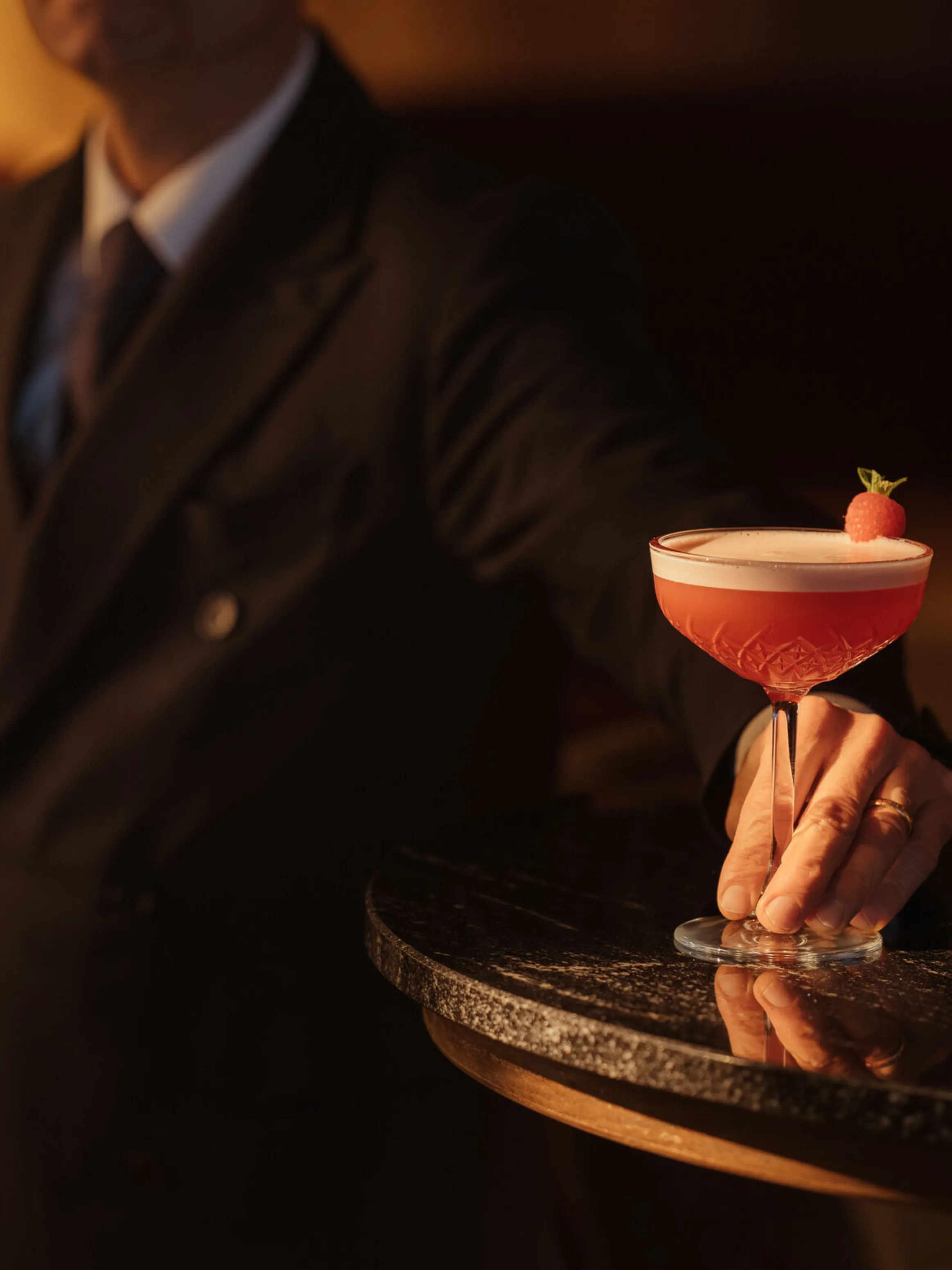A bartender in a suit presents a stylish pink cocktail garnished with a mint leaf on a dark, reflective table. The setting is warm and elegant.