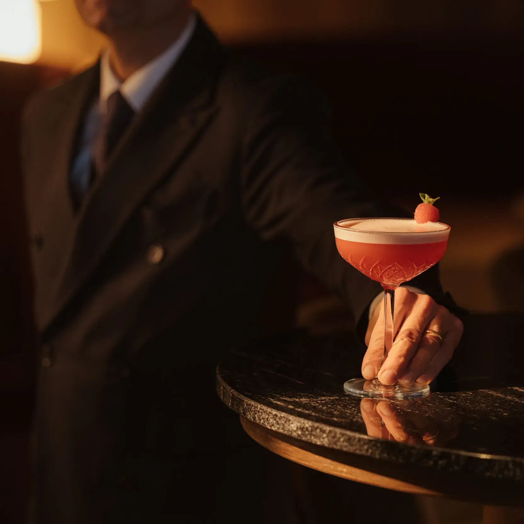 A bartender in a suit presents a stylish pink cocktail garnished with a mint leaf on a dark, reflective table. The setting is warm and elegant.