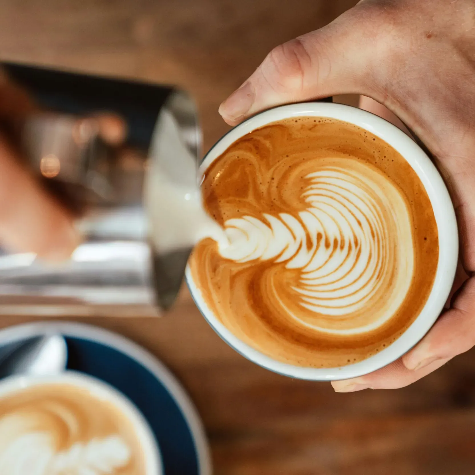 Barista’s hands pouring cappuccino foam into a cup; in the background, on a wooden counter, a second cappuccino ready to be enjoyed hot.