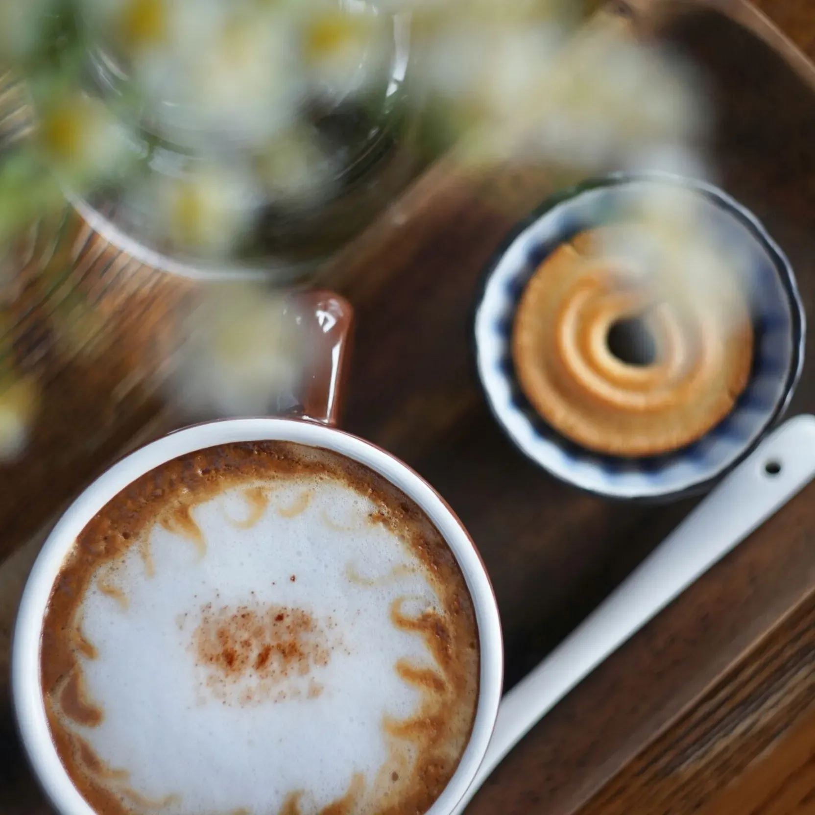 Cappuccino with a dry pastry cookie, served on a small wooden tray; next to it, a slightly blurred vase of daisies, viewed from above.