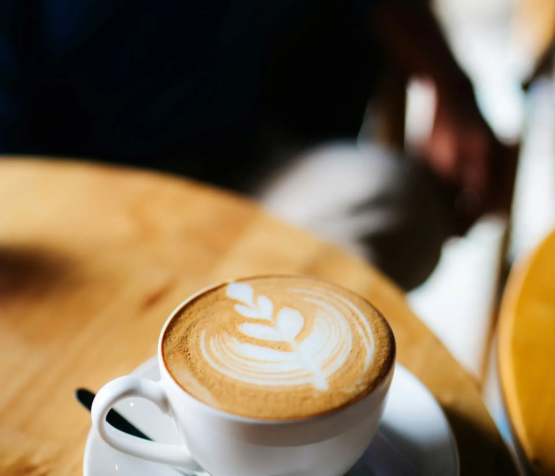 Cappuccino in a white cup on a white saucer with a teaspoon, resting on a wooden table; in the background, a blurred seated person.