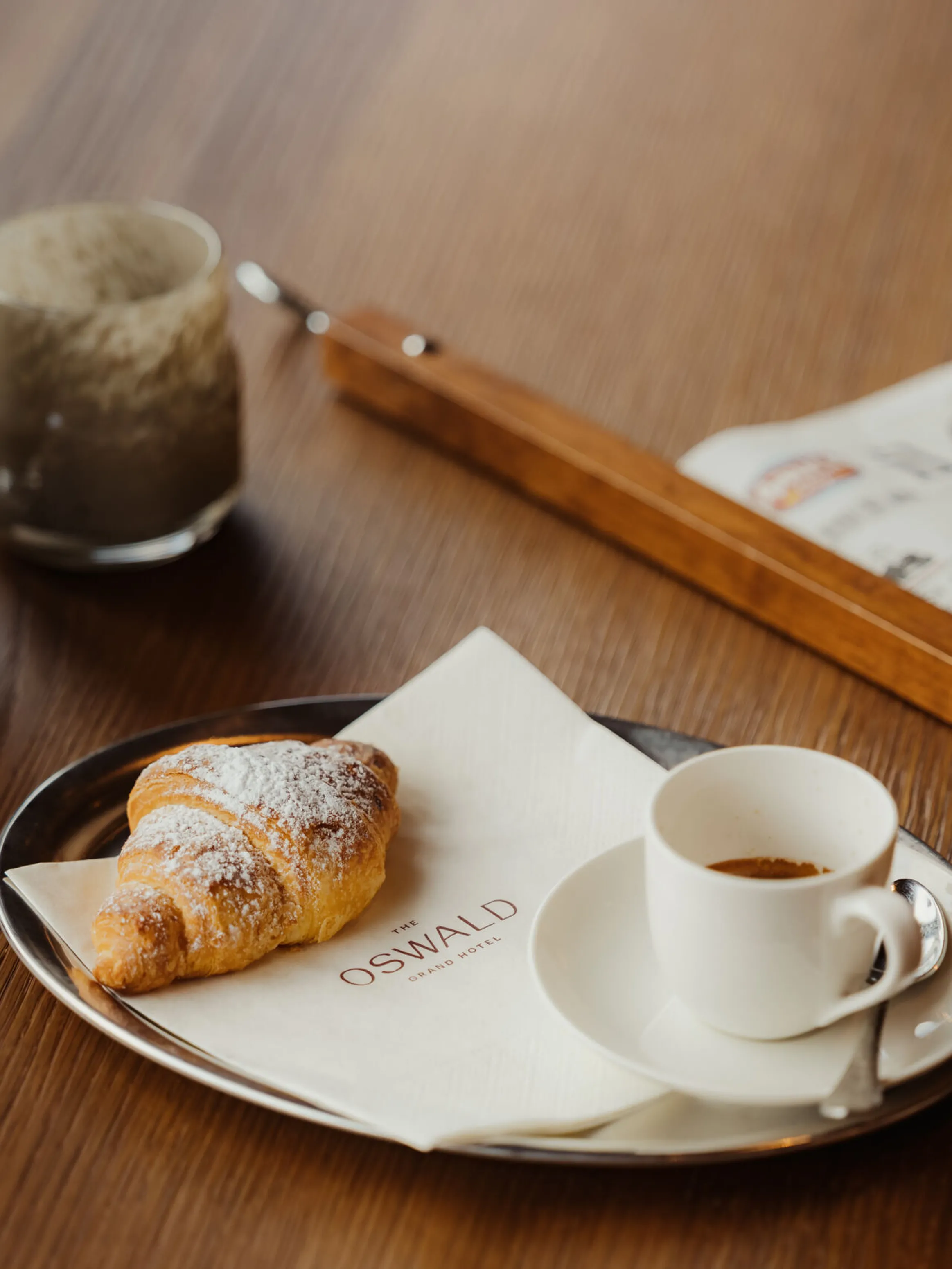 A croissant on a napkin with a cup of espresso rests on a tray on a wooden table. A newspaper and decorative vases nearby add a cozy atmosphere.
