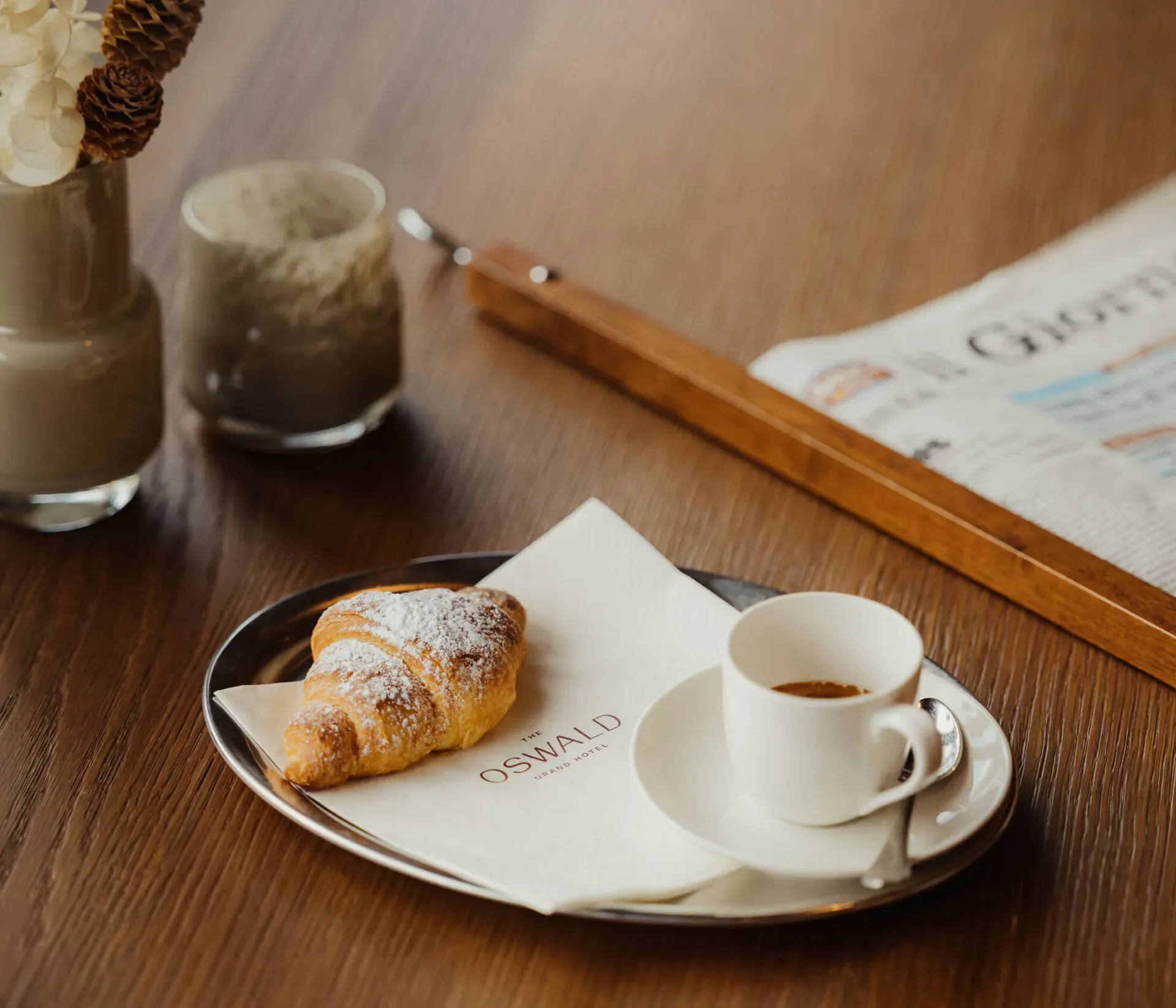 A croissant on a napkin with a cup of espresso rests on a tray on a wooden table. A newspaper and decorative vases nearby add a cozy atmosphere.