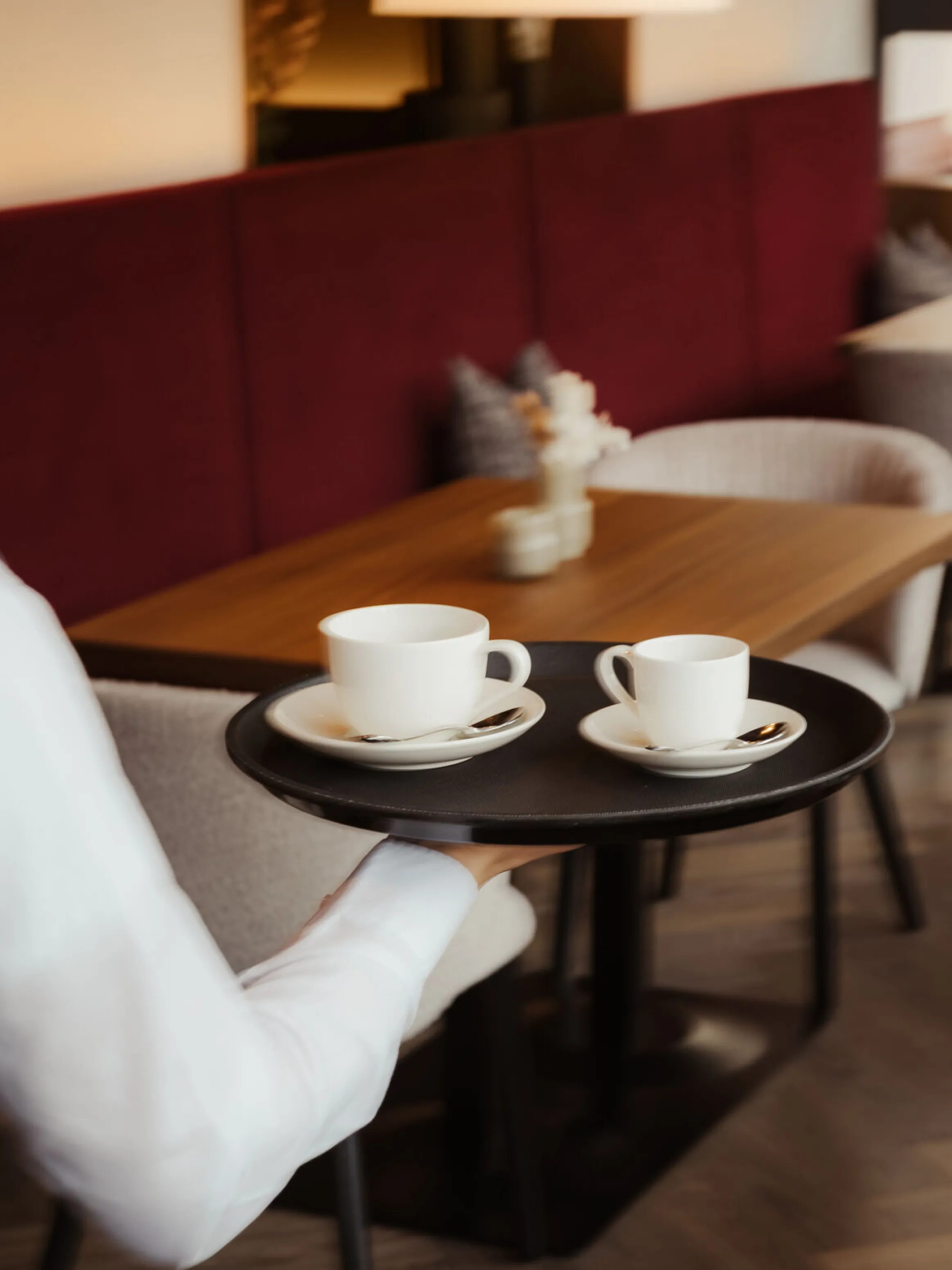 A waiter carries a tray with two coffee cups in a cozy café. The setting features plush red seating, wooden tables, and a calm ambiance.