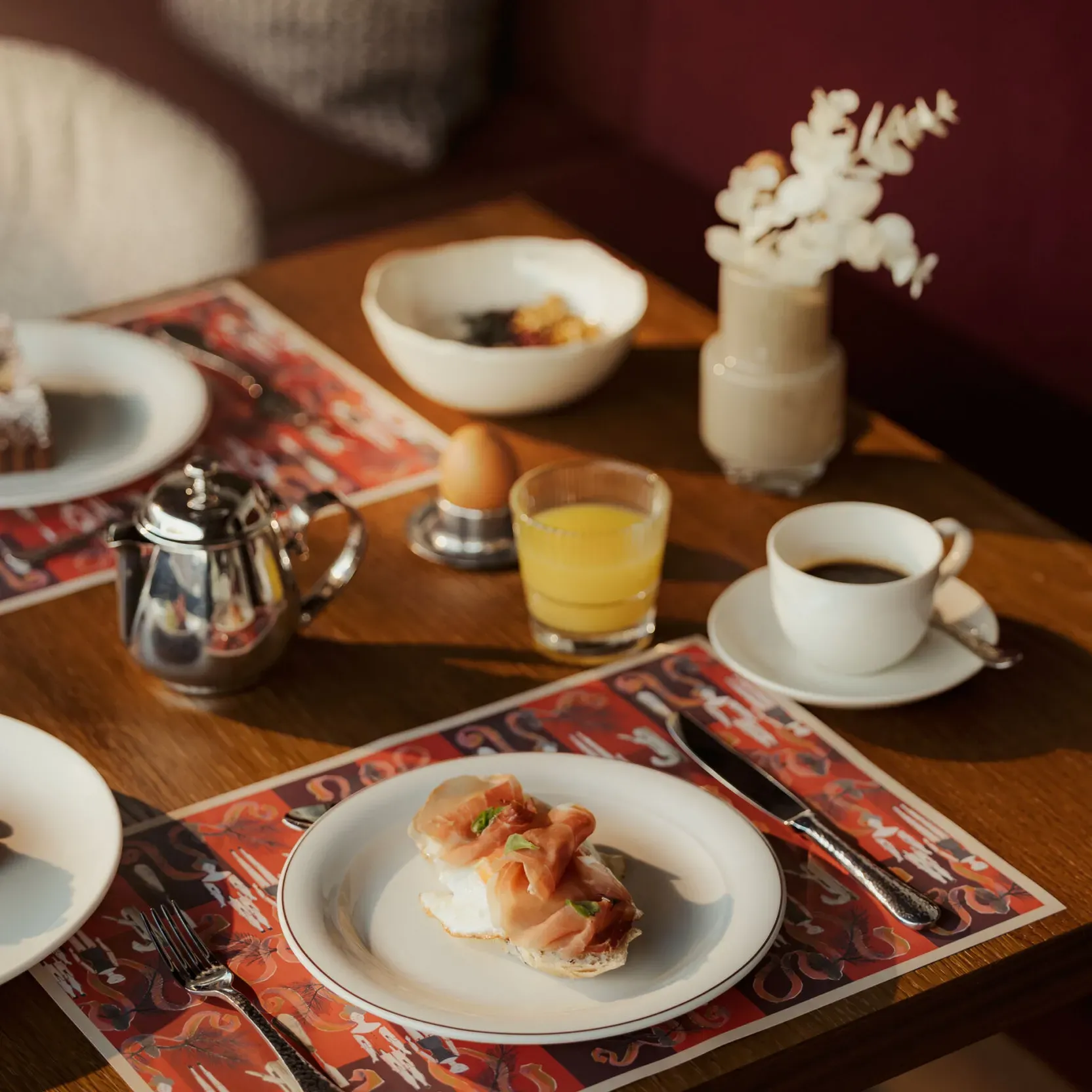Breakfast setting with toast topped with smoked salmon and greens. Nearby are a teapot, boiled egg, orange juice, coffee cup, and flower vase. Cozy and inviting.