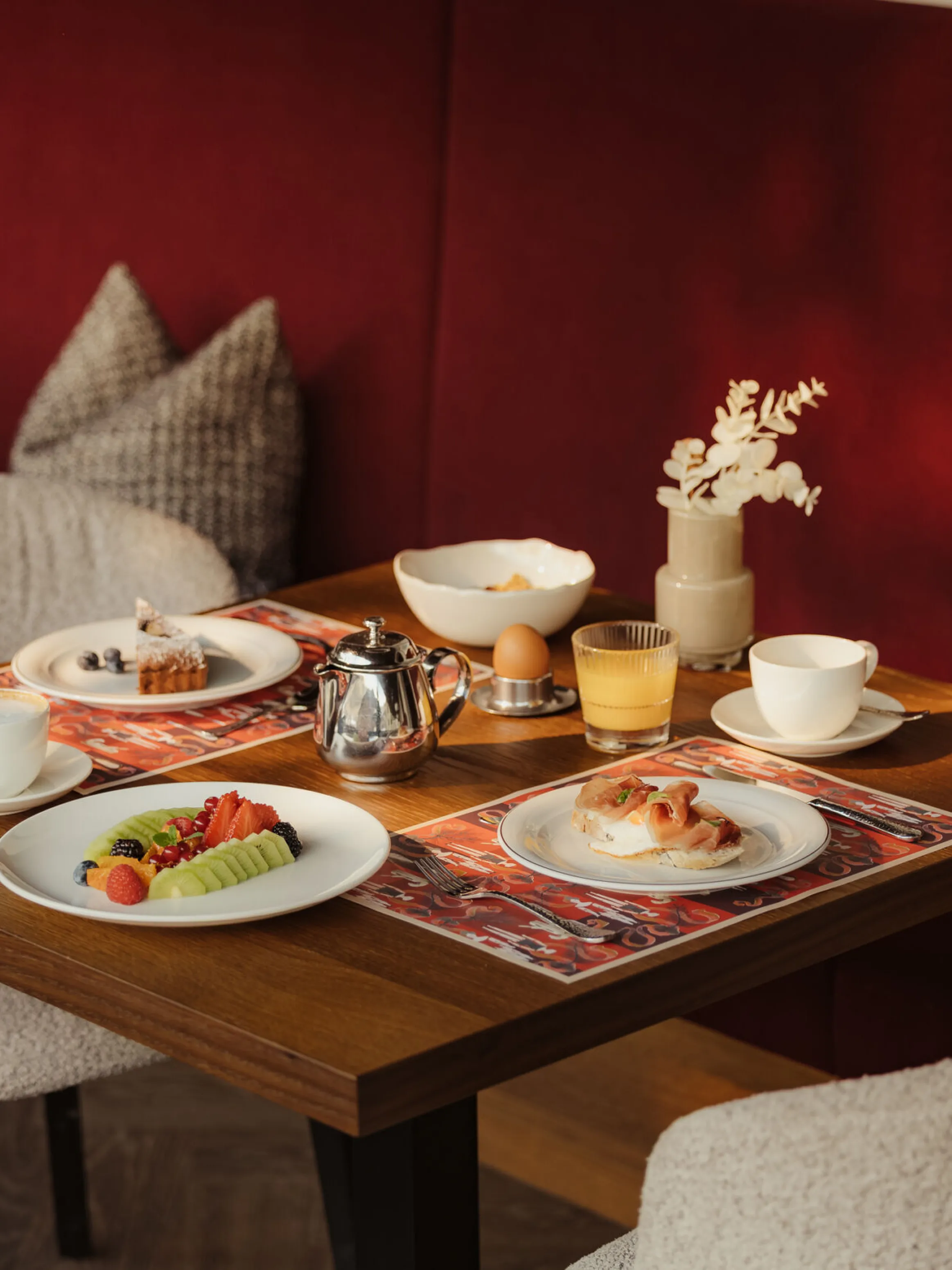 Cozy breakfast scene with two plates of pastries, fruit, and tea on a sunlit wooden table. Red cushioned bench and elegant decor add warmth.