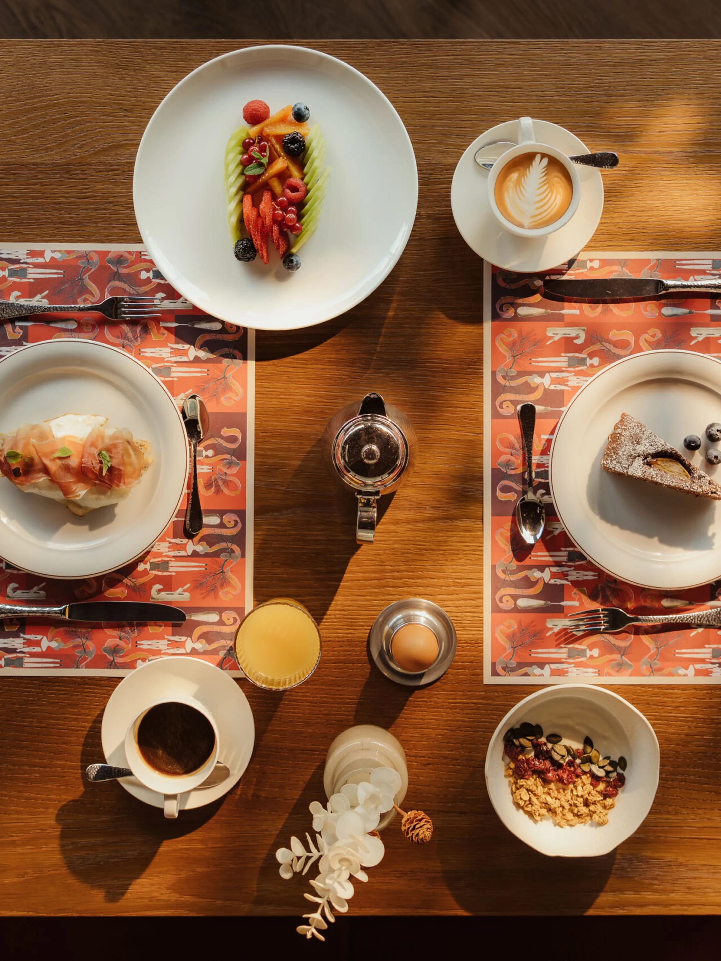 A cozy breakfast table set for two with various dishes, coffee, and juice. The mood is warm and inviting, with a dessert, latte, and croissant.