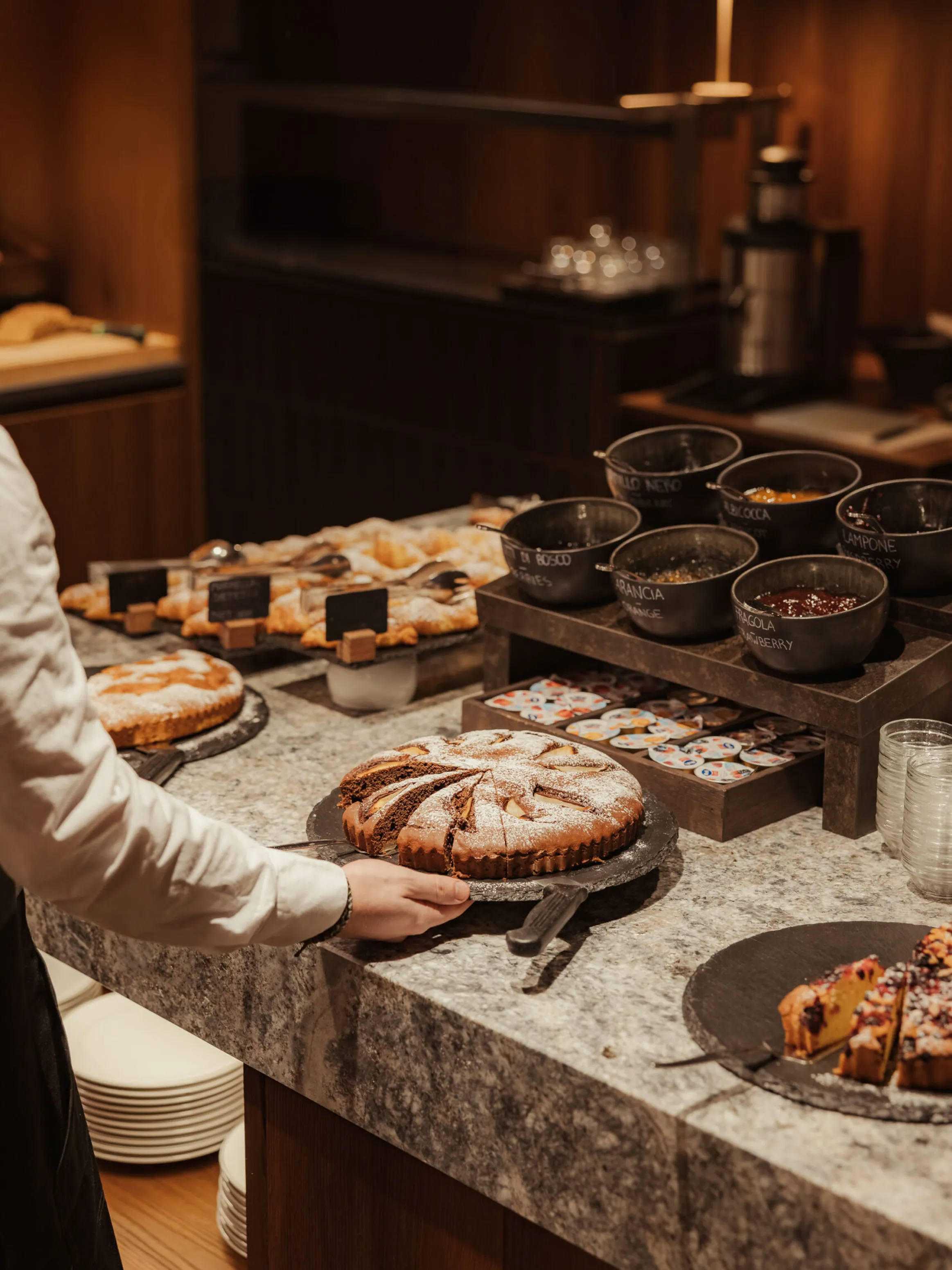 A person’s hand reaches for a powdered sugar-topped cake on a marble counter. Nearby are pastries, bowls of spreads, and a cozy, warm-lit ambiance.