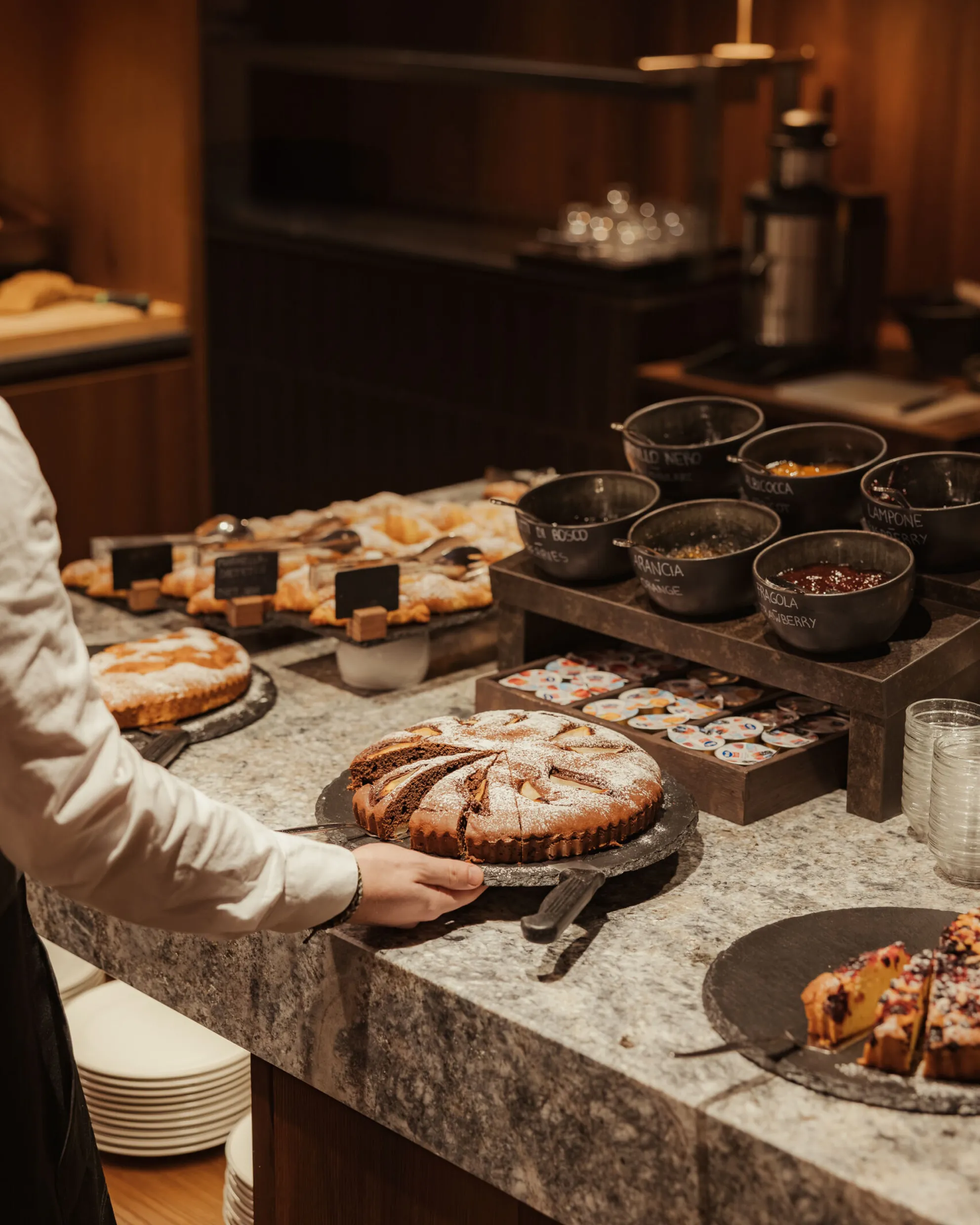 A person’s hand reaches for a powdered sugar-topped cake on a marble counter. Nearby are pastries, bowls of spreads, and a cozy, warm-lit ambiance.