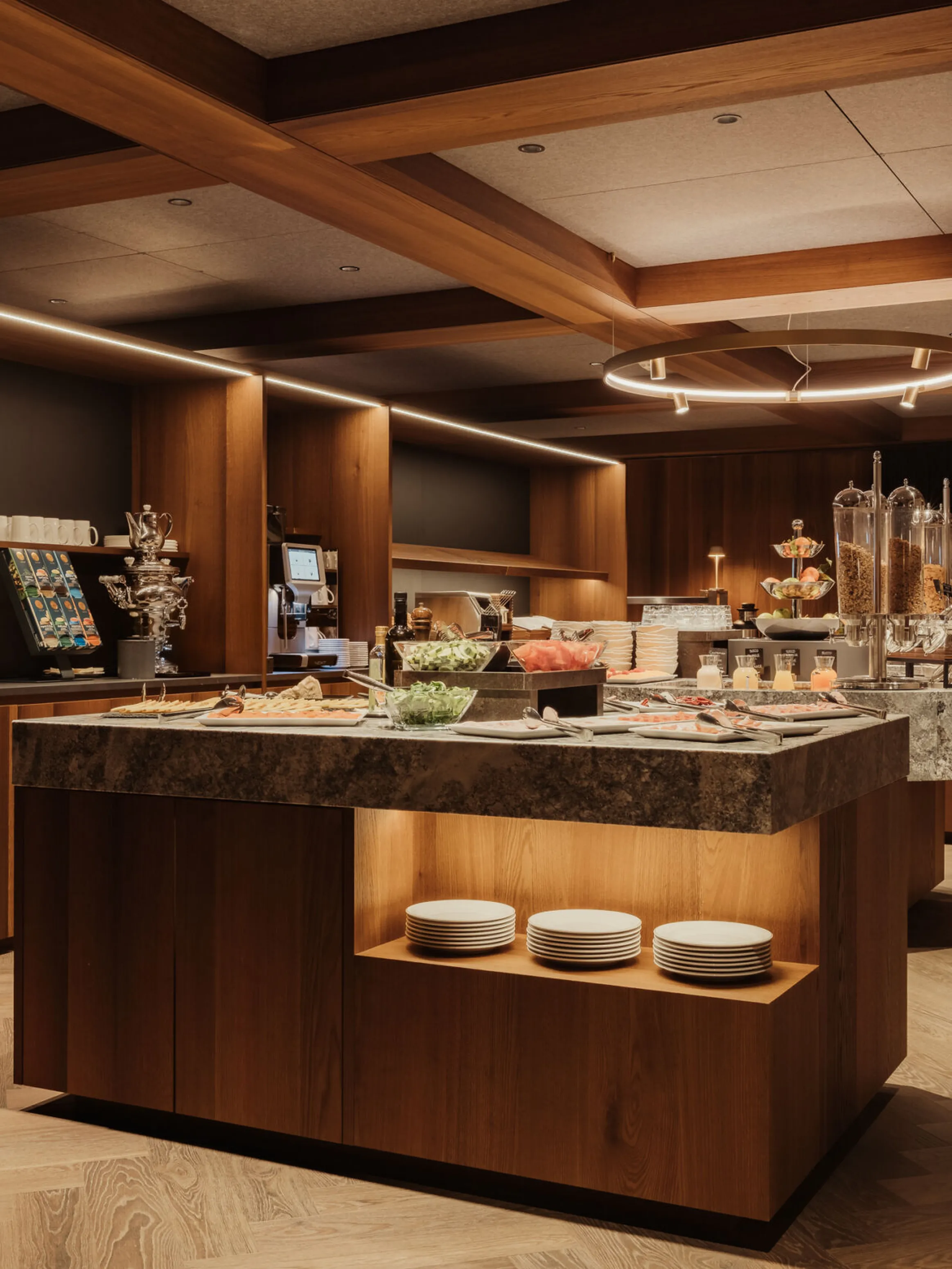 A modern buffet area with warm wood accents, offering pastries, fruits, and cereal dispensers. Elegant lighting and neatly arranged plates create a cozy, inviting atmosphere.