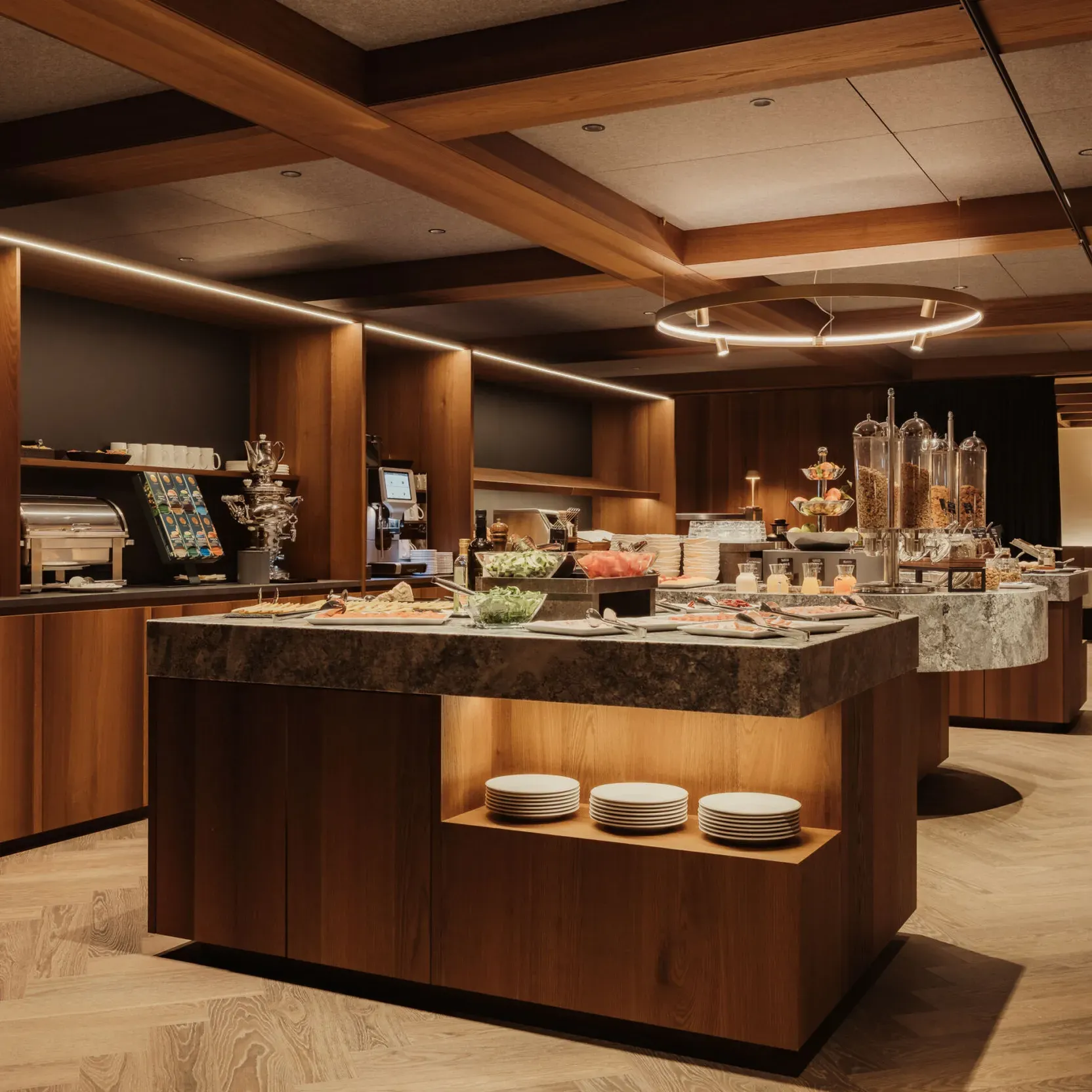A modern buffet area with warm wood accents, offering pastries, fruits, and cereal dispensers. Elegant lighting and neatly arranged plates create a cozy, inviting atmosphere.
