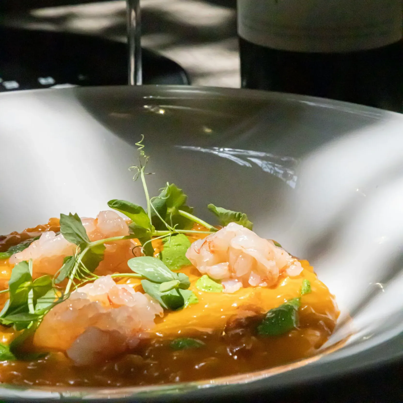 Close-up of a gourmet shrimp dish, accompanied by sauce and decorative leaves; in the background, a bottle of wine and a glass of white wine.