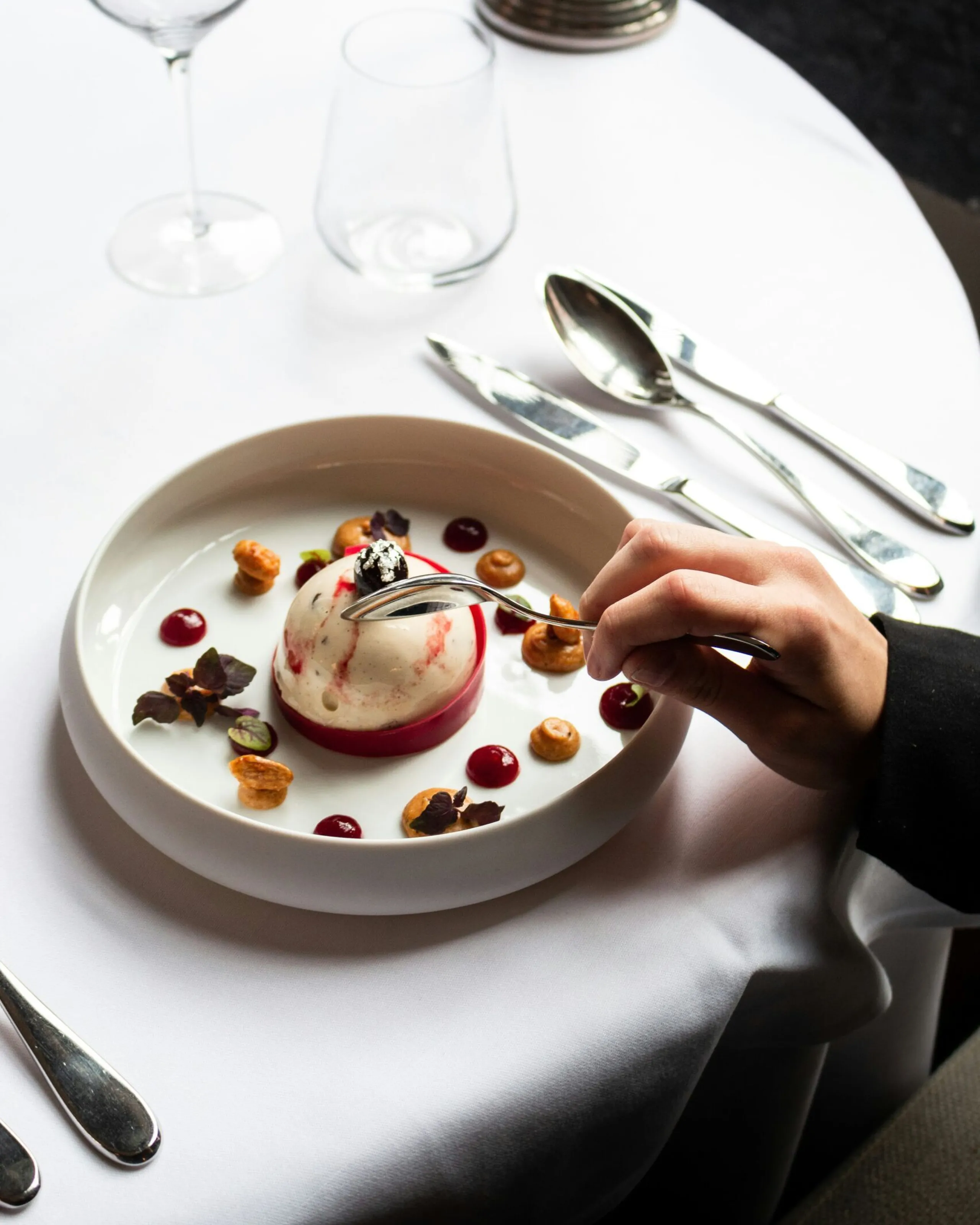 Single-serving tart with white chocolate mousse on a strawberry shortcrust base, ready to be tasted by a guest holding a spoon; the plate is decorated with drops of red fruit sauce and placed on a white cotton tablecloth covering a round table.
