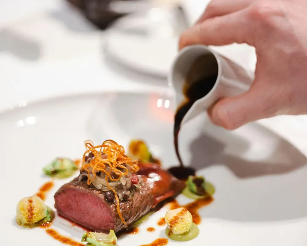 Beef fillet served on a white plate, elegantly plated with fried julienned carrots, while the waiter’s hand pours the accompanying sauce; in the background, a bread basket and a glass of red wine.