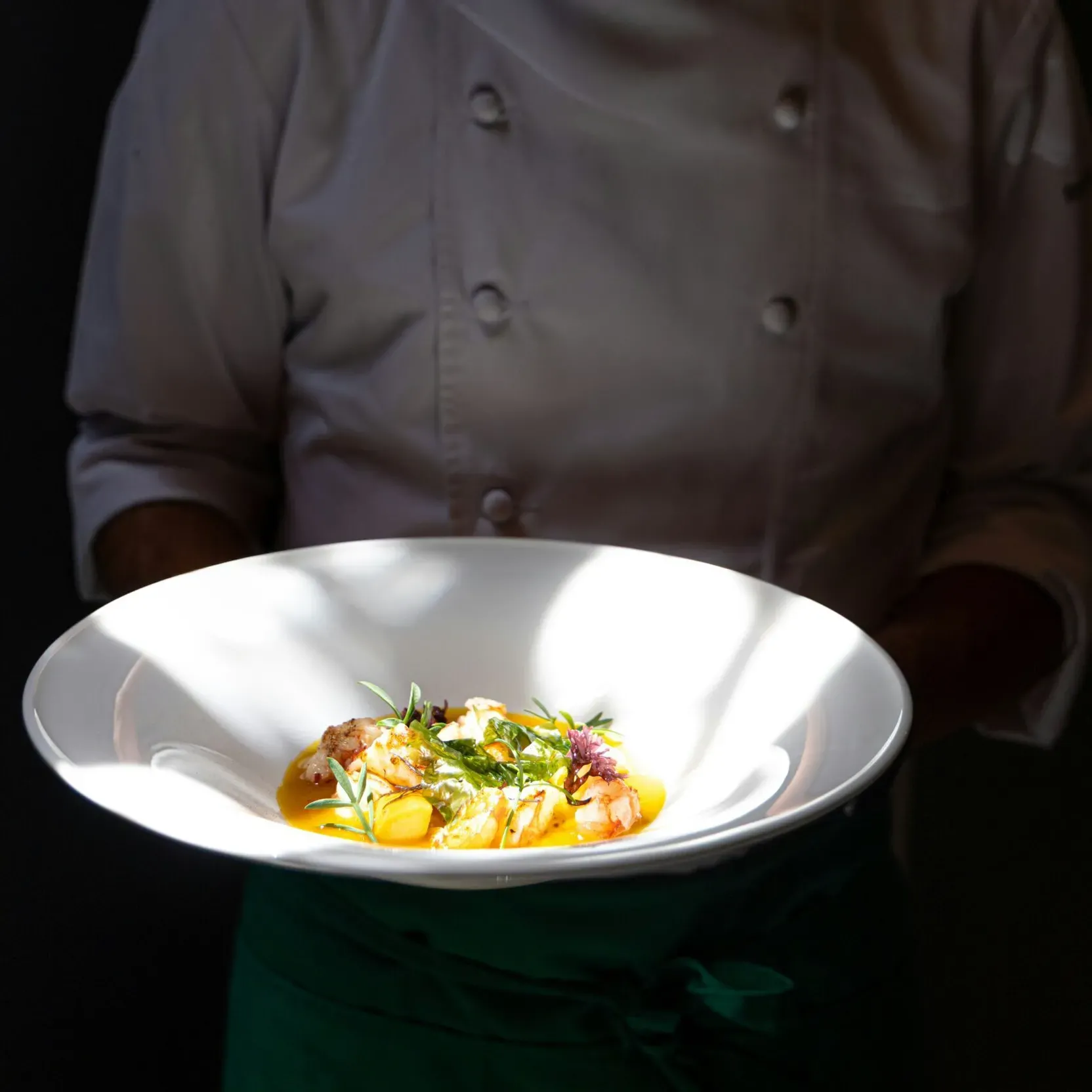 Chef in dim light holding a freshly prepared gourmet plate.
