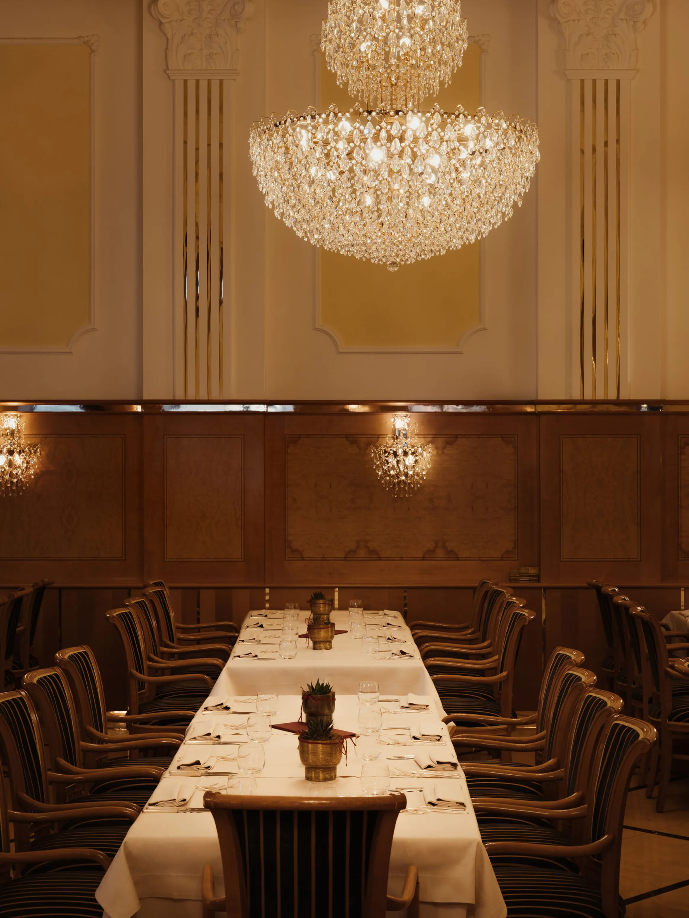 Elegant dining room with a long, white-clothed table surrounded by wooden chairs. Large crystal chandeliers hang above, creating a warm and luxurious atmosphere.