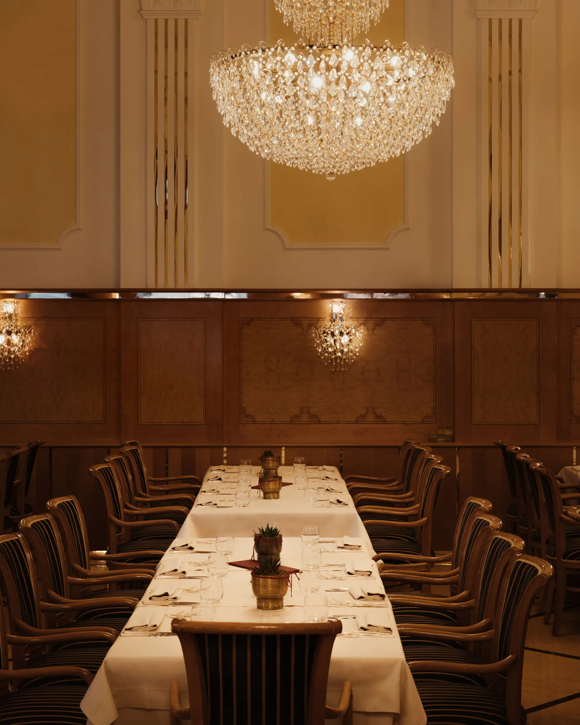 Elegant dining room with a long, white-clothed table surrounded by wooden chairs. Large crystal chandeliers hang above, creating a warm and luxurious atmosphere.