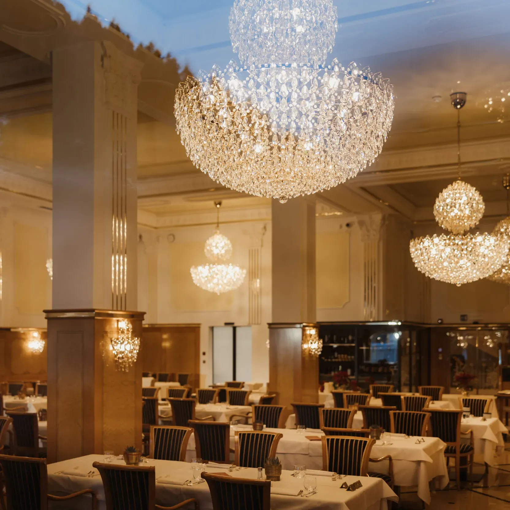 Elegant dining room with sparkling chandeliers, neatly arranged tables with white tablecloths, and a warm, inviting ambiance exuding luxury and sophistication.