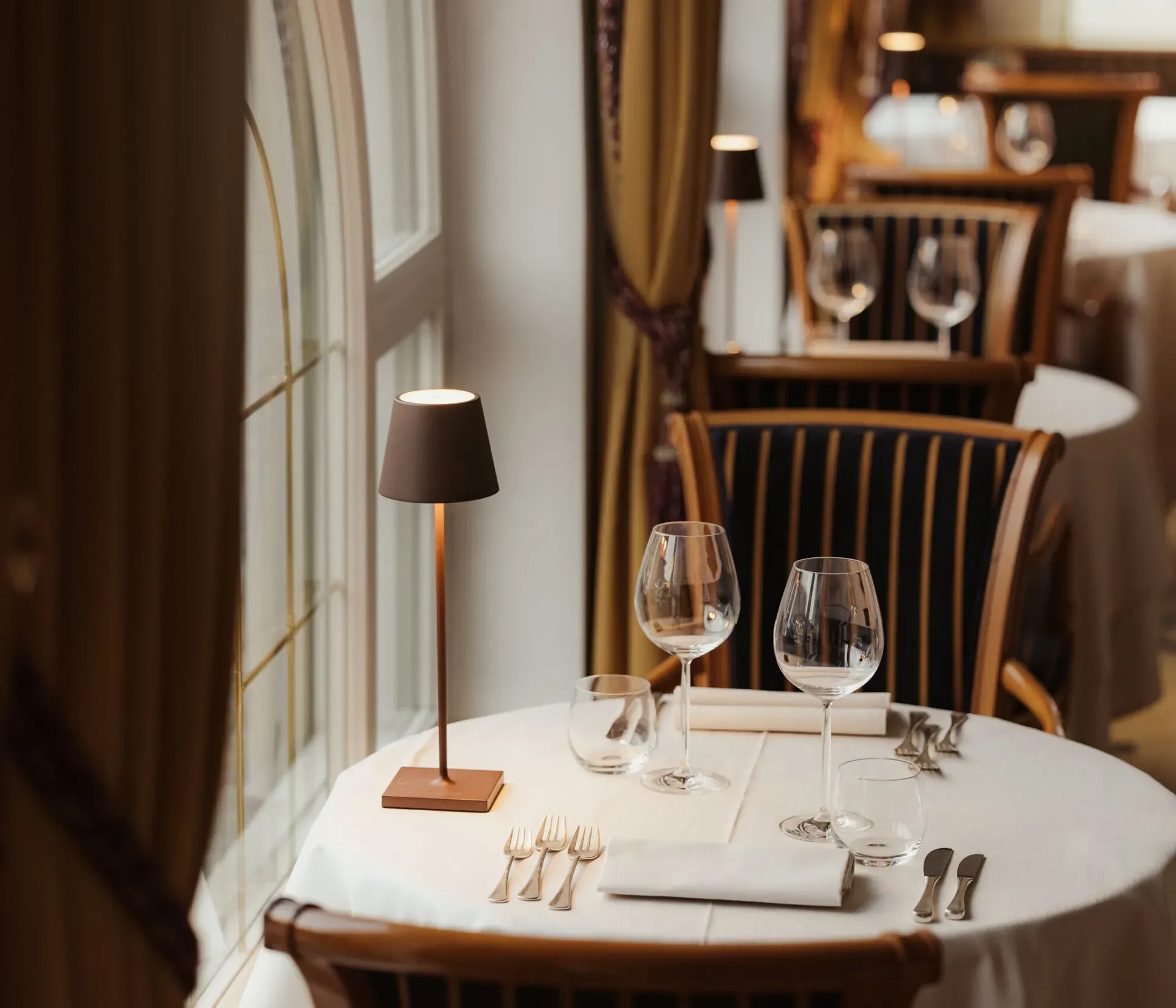 Elegant restaurant interior with a round table set for two, featuring wine glasses, cutlery, and a small table lamp. The atmosphere is cozy and intimate.