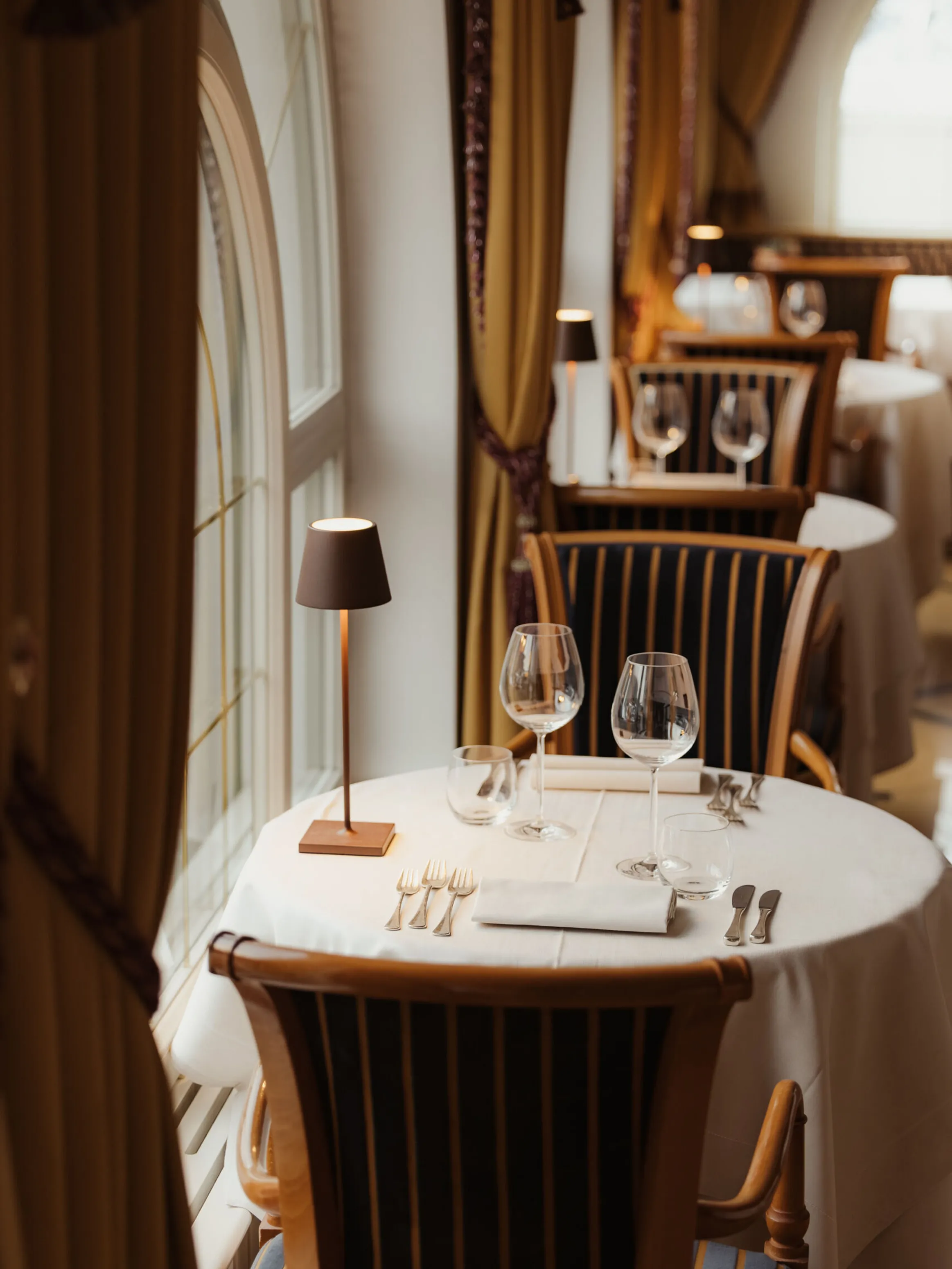 Elegant restaurant setting with round tables, white tablecloths, and wooden chairs. Soft lighting from small lamps creates a cozy, intimate ambiance.