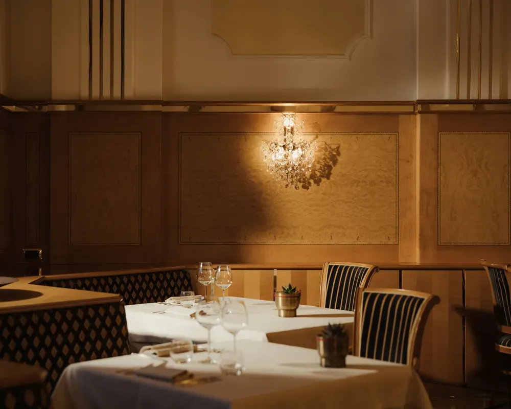 Elegant restaurant with dim lighting, featuring a crystal chandelier, wooden paneling, and neatly set tables with striped chairs, creating a cozy atmosphere.