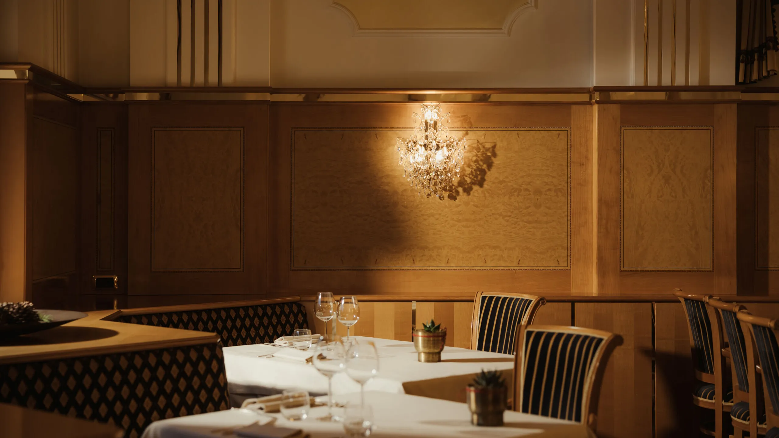 Elegant restaurant with dim lighting, featuring a crystal chandelier, wooden paneling, and neatly set tables with striped chairs, creating a cozy atmosphere.