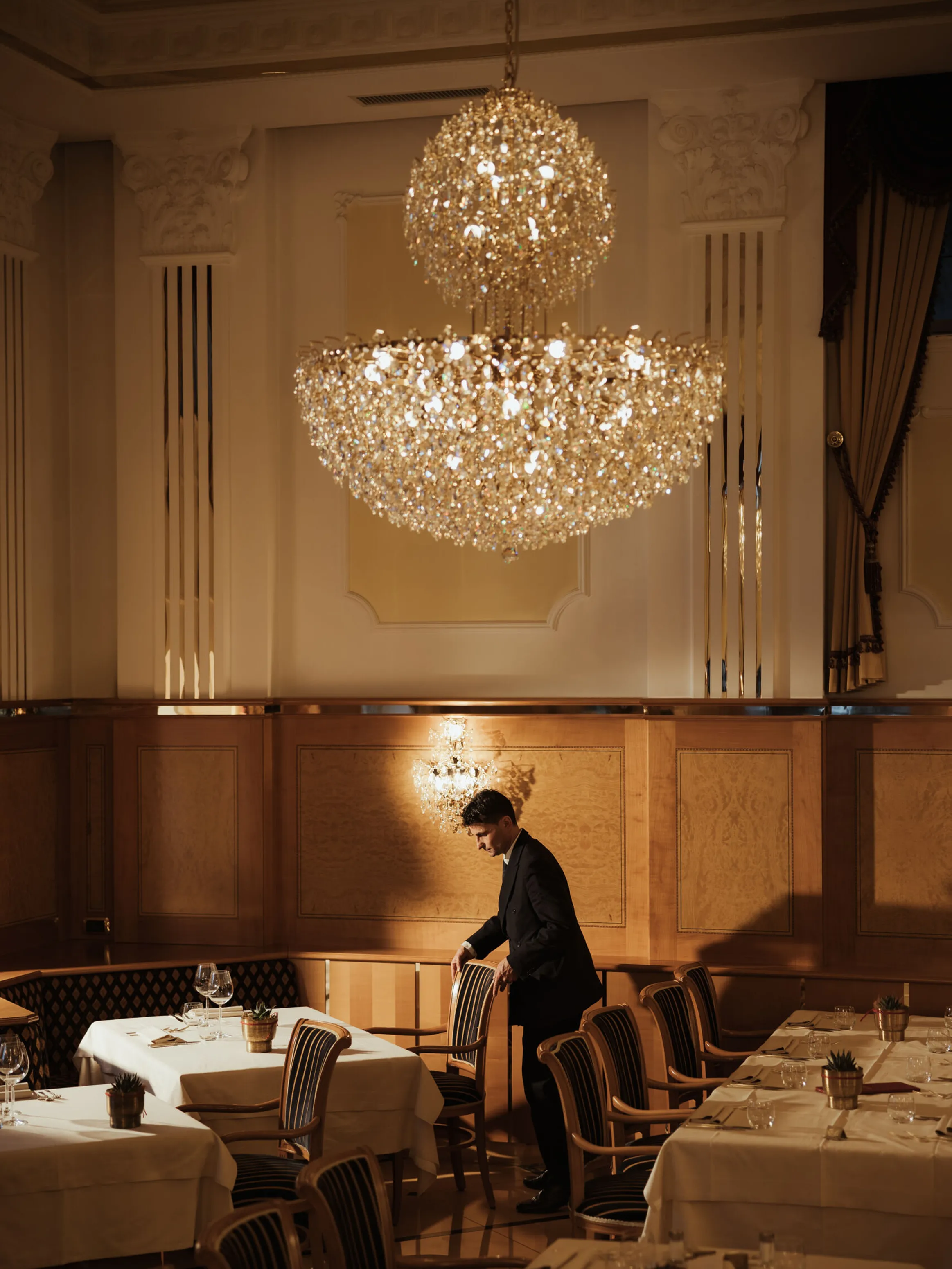 Elegant dining room with a grand chandelier, white tablecloths, and wooden chairs. A waiter adjusts a chair, creating a refined, warm atmosphere.