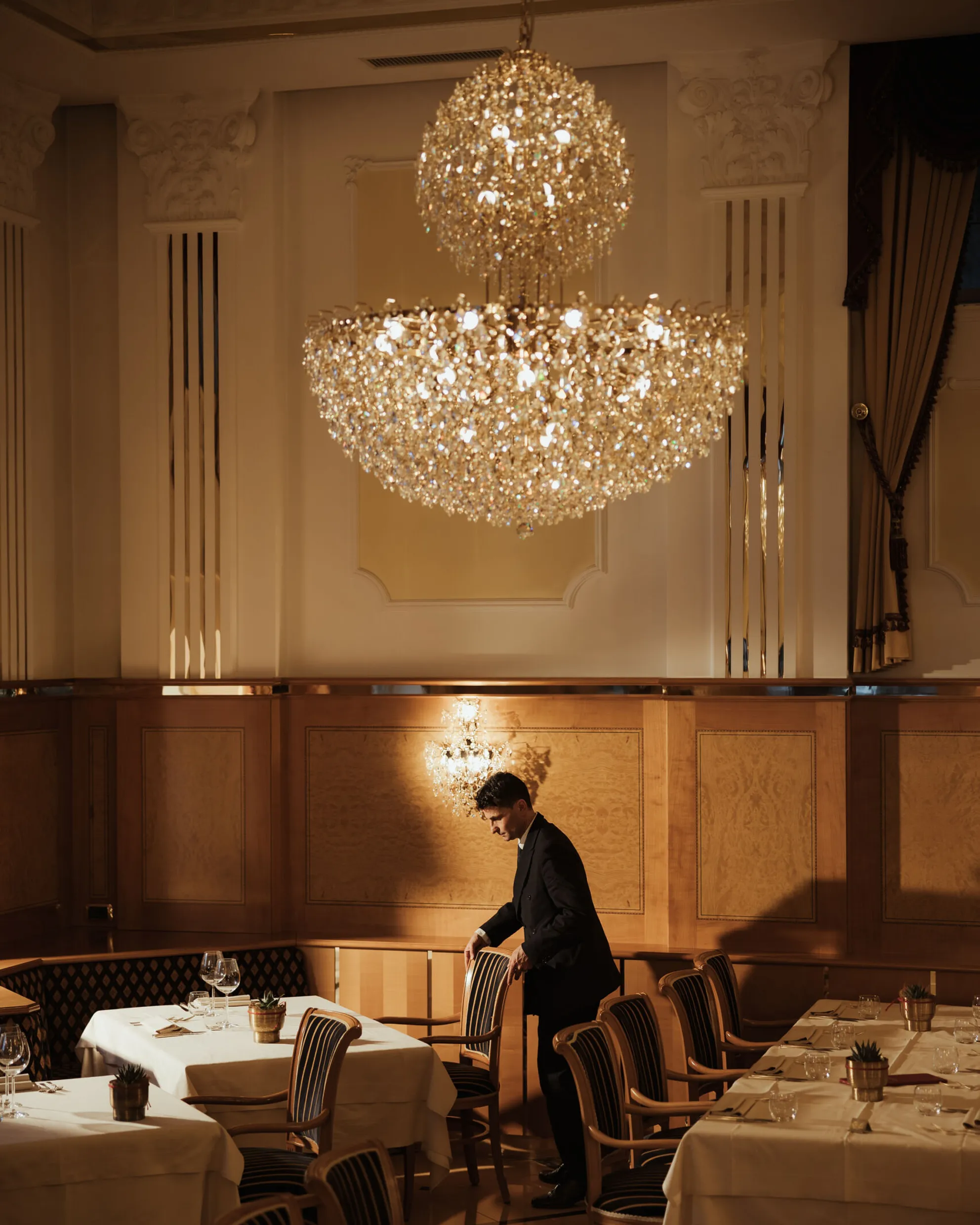 Elegant dining room with a grand chandelier, white tablecloths, and wooden chairs. A waiter adjusts a chair, creating a refined, warm atmosphere.