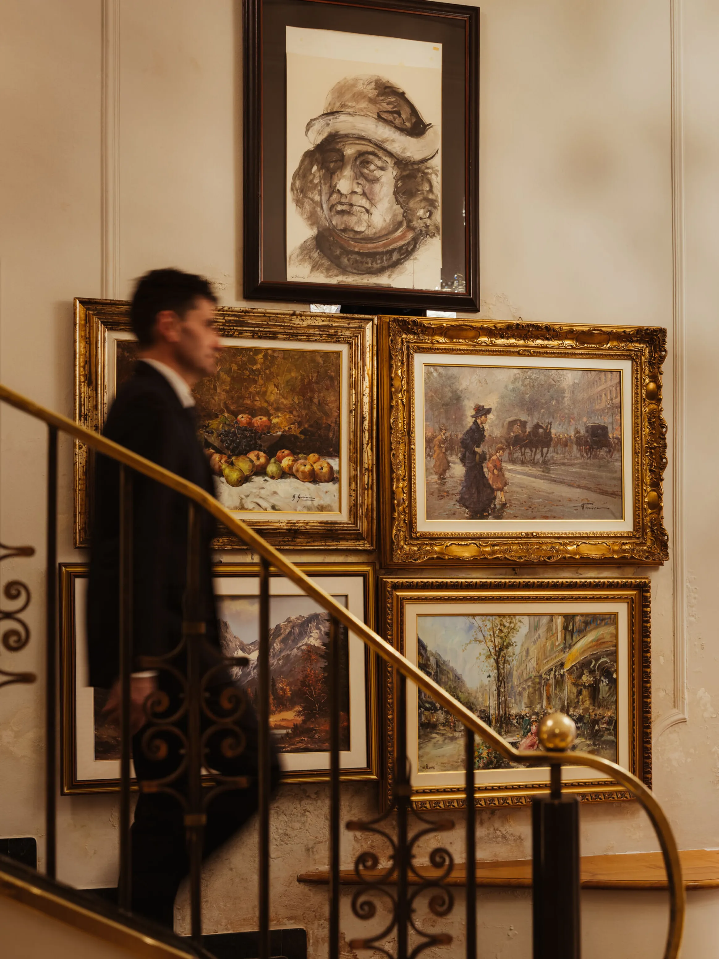 A man in a suit ascends a curved staircase adorned with ornate railings. Behind him, framed paintings depict various scenes, adding an elegant, artistic ambiance.
