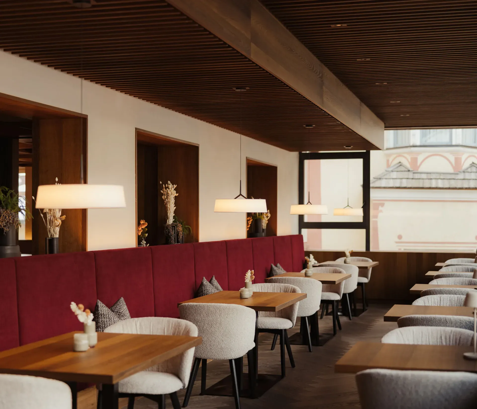 A cozy restaurant interior with wooden tables, soft white chairs, and a red cushioned wall. Modern pendant lights hang above, creating a warm atmosphere.
