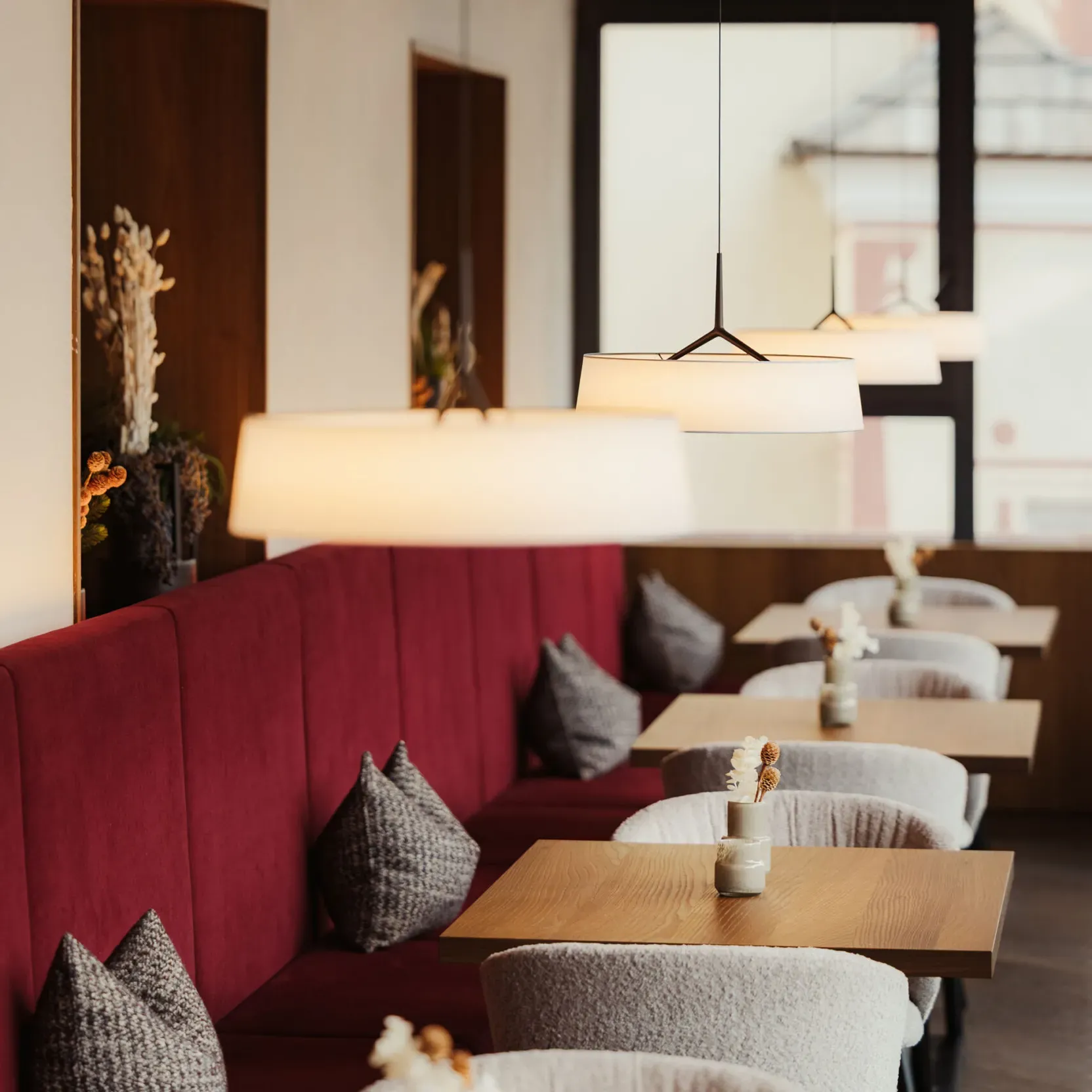 Modern cafe interior with red booth seating, gray pillows, wooden tables, and white pendant lights. Minimalist decor includes small vases with dried flowers, creating a cozy, inviting atmosphere.