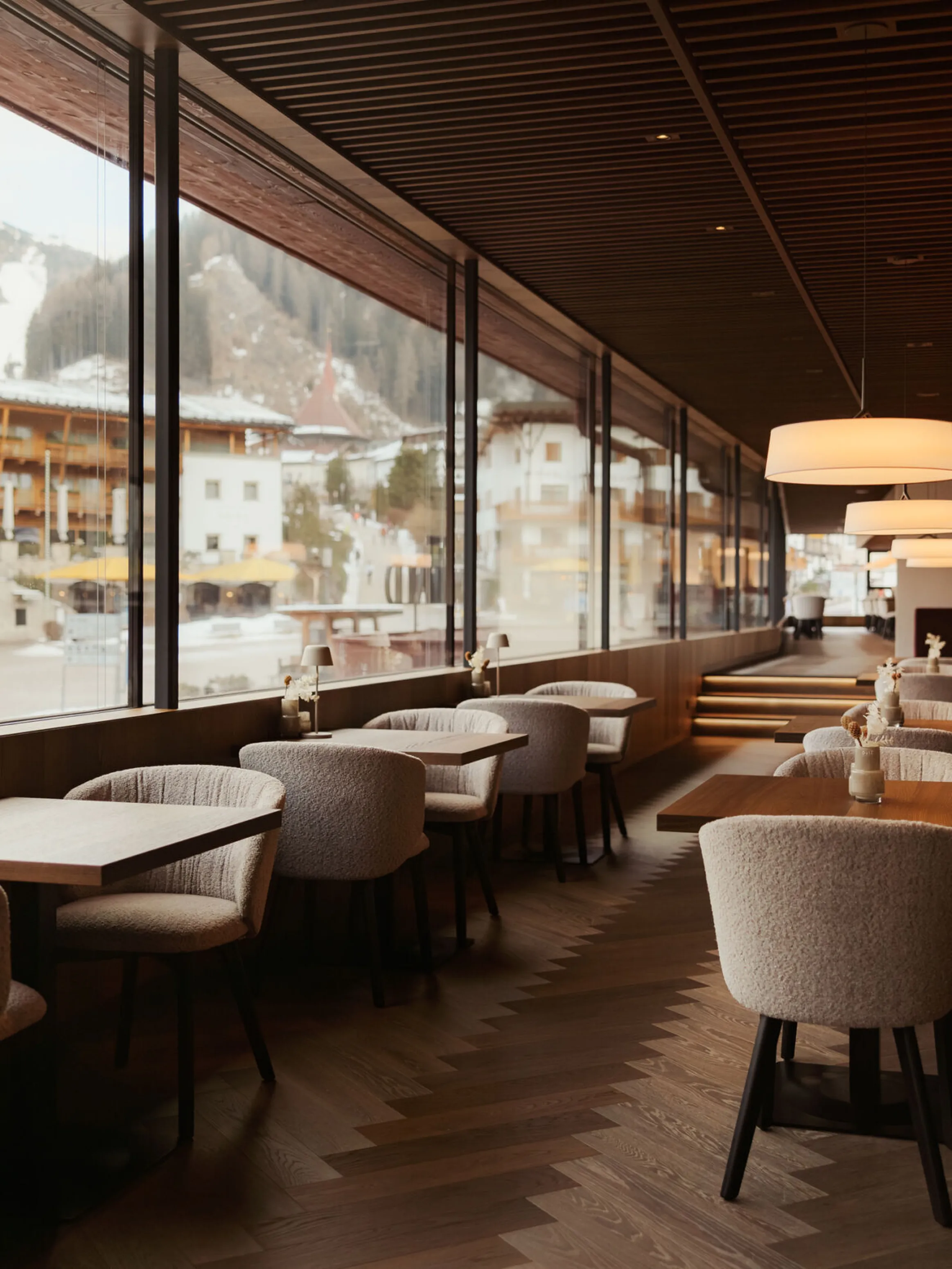 Cozy restaurant interior with wooden floors, plush gray chairs, and maroon cushioned seating. Large windows showcase a snowy alpine village view.