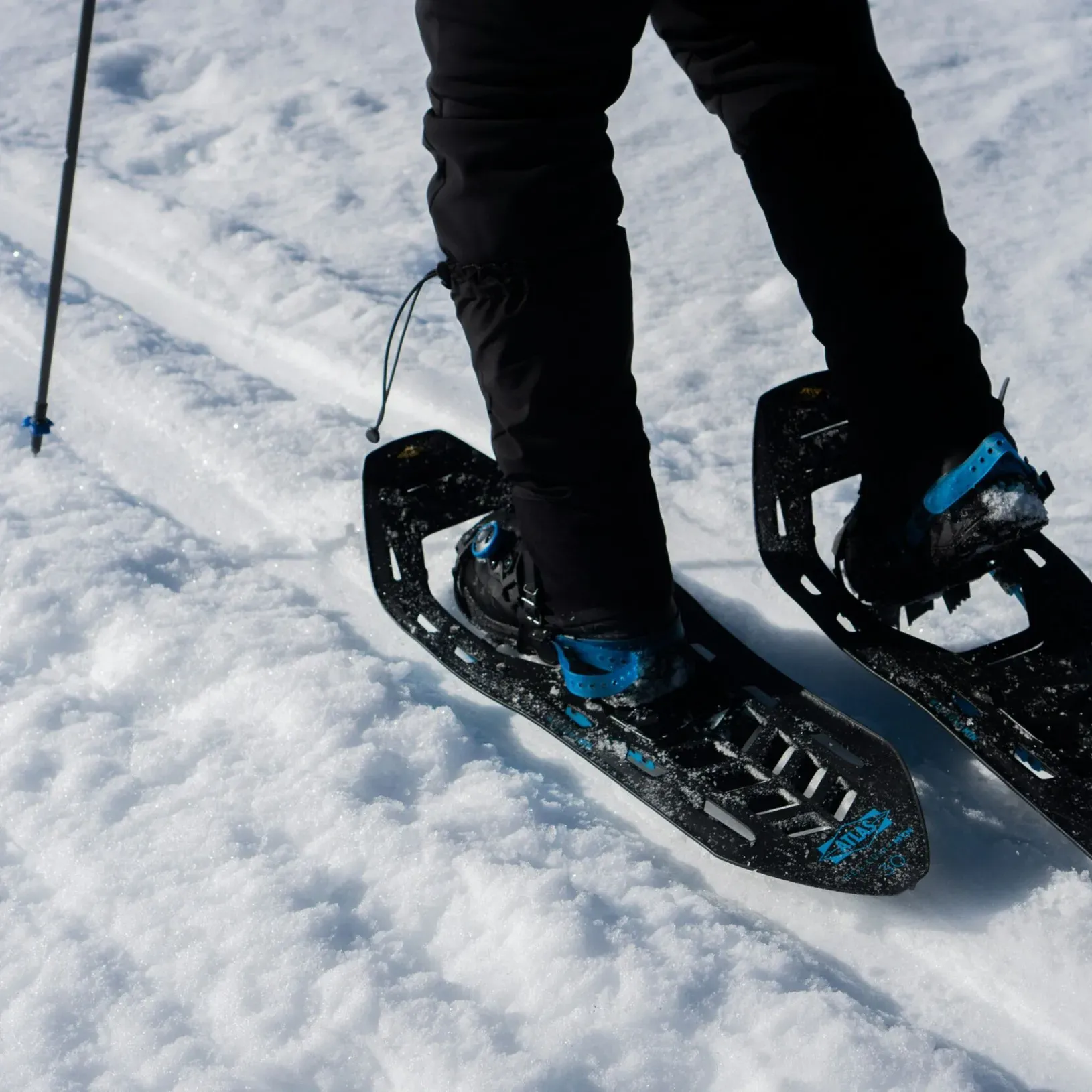 Close-up of feet walking on packed snow along a winter trail, wearing snowshoes and using trekking poles.