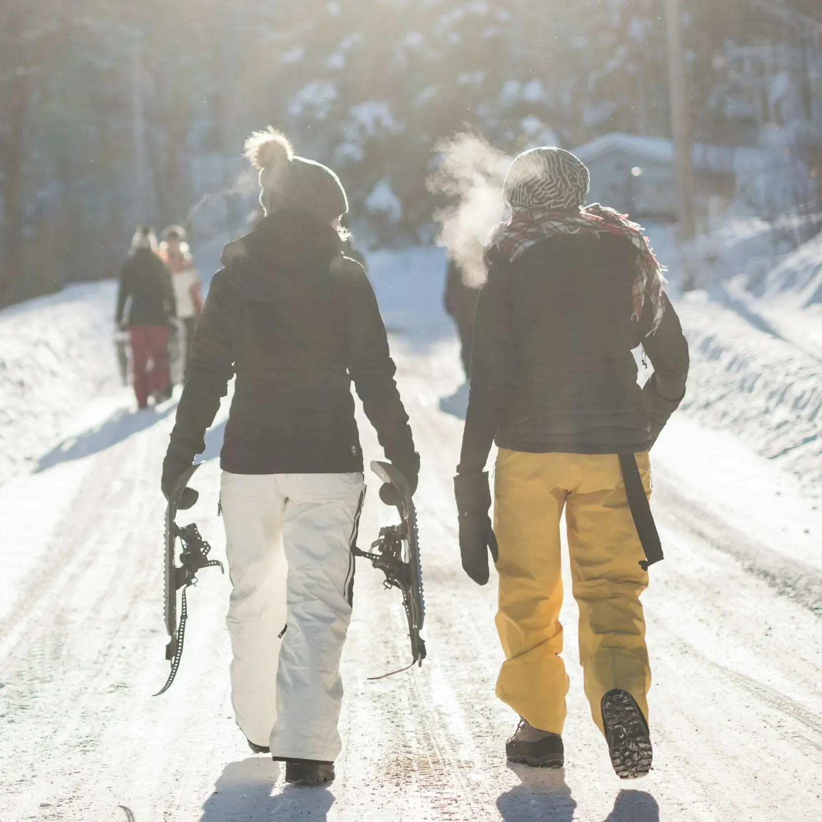 Two friends in winter outdoor gear chatting as they prepare to start a hike on fresh snow, holding snowshoes, their breath visible in the cold mountain air, with other people in the background along the snowy trail.