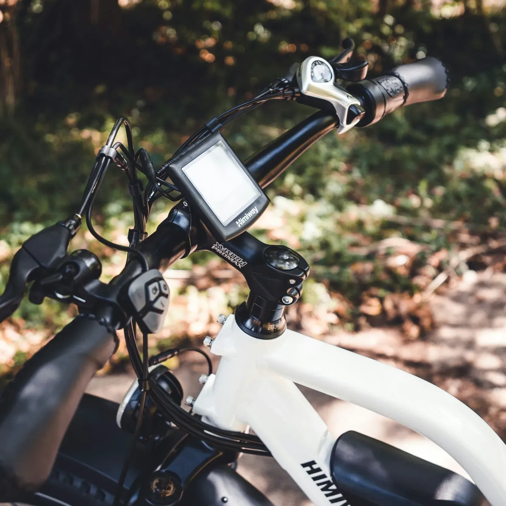 Close-up of the handlebar of a white e-bike with onboard computer, parked in a forest clearing on a sunny day.