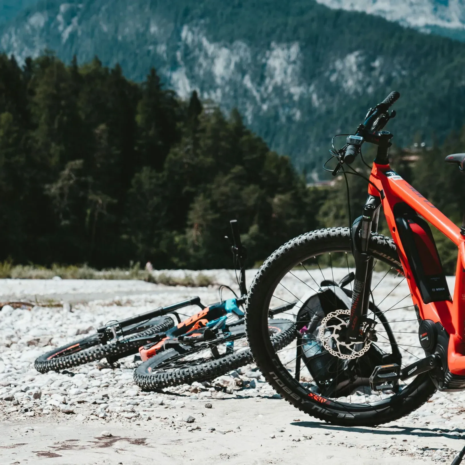 Two e-bikes parked by a mountain stream during a summer hike break, one on its kickstand and the other lying down, with mountain cliffs and a spruce forest framing the background on a warm, sunny day.