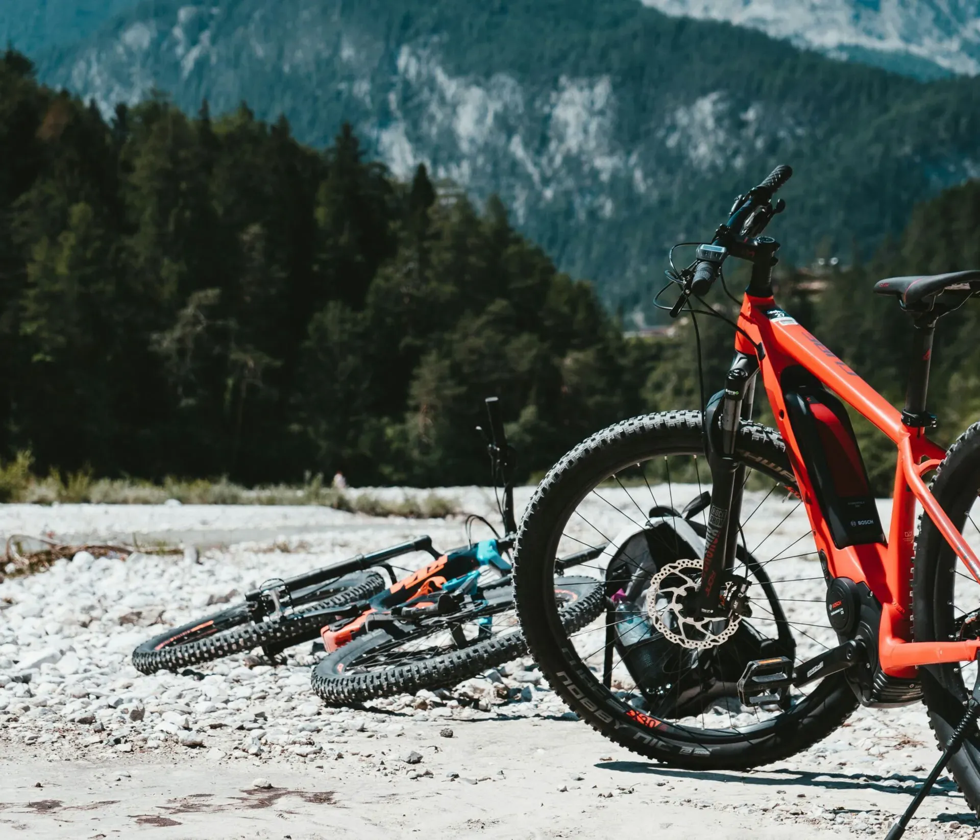 Two e-bikes parked by a mountain stream during a summer hike break, one on its kickstand and the other lying down, with mountain cliffs and a spruce forest framing the background on a warm, sunny day.