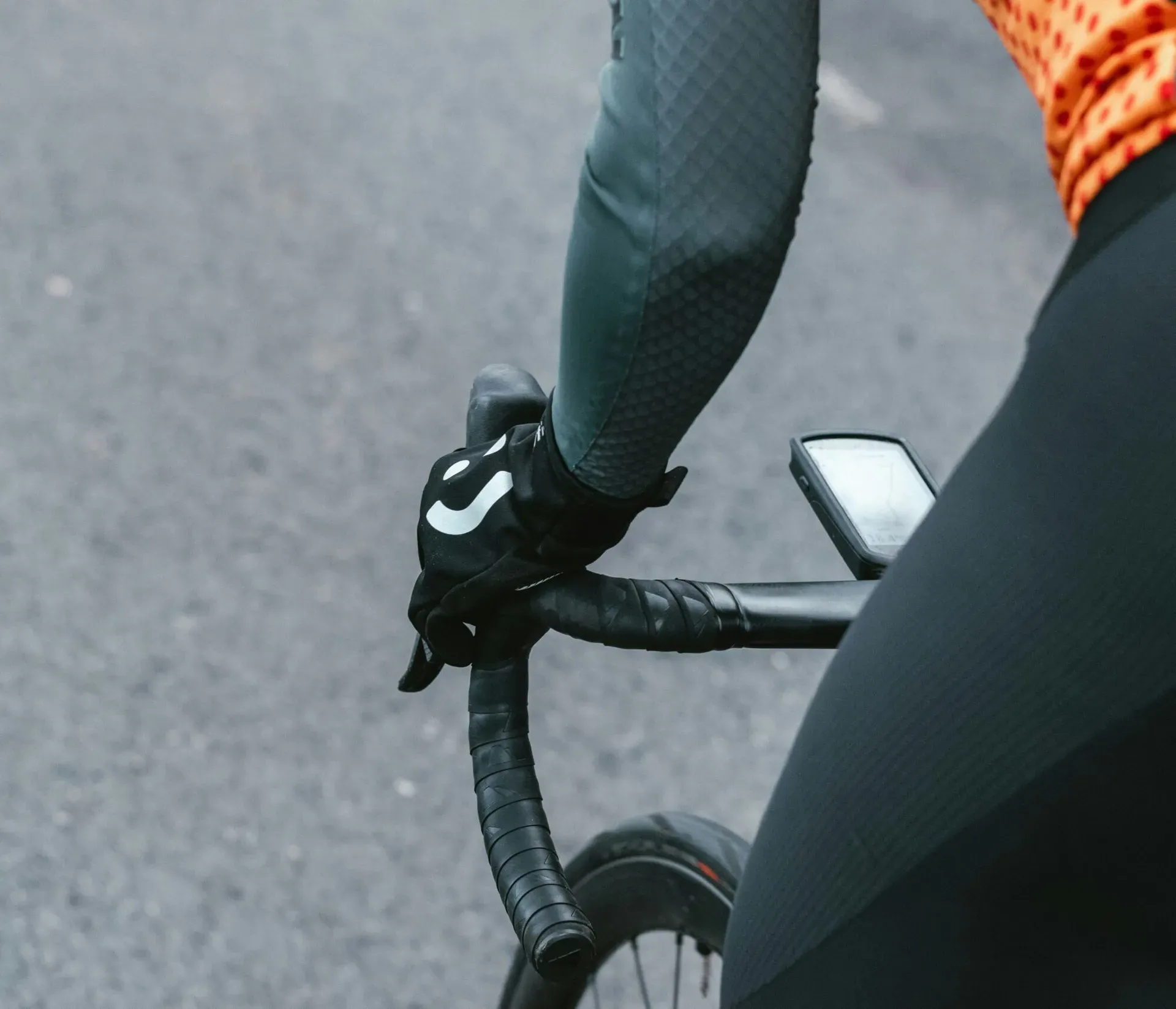 Close-up of a road bike on the road with a cyclist pedaling standing up, wearing gloves, arm warmers, an orange t-shirt, and shorts; showing the road surface, front wheel, handlebar with onboard computer, and the cyclist’s hand and hip while pedaling.