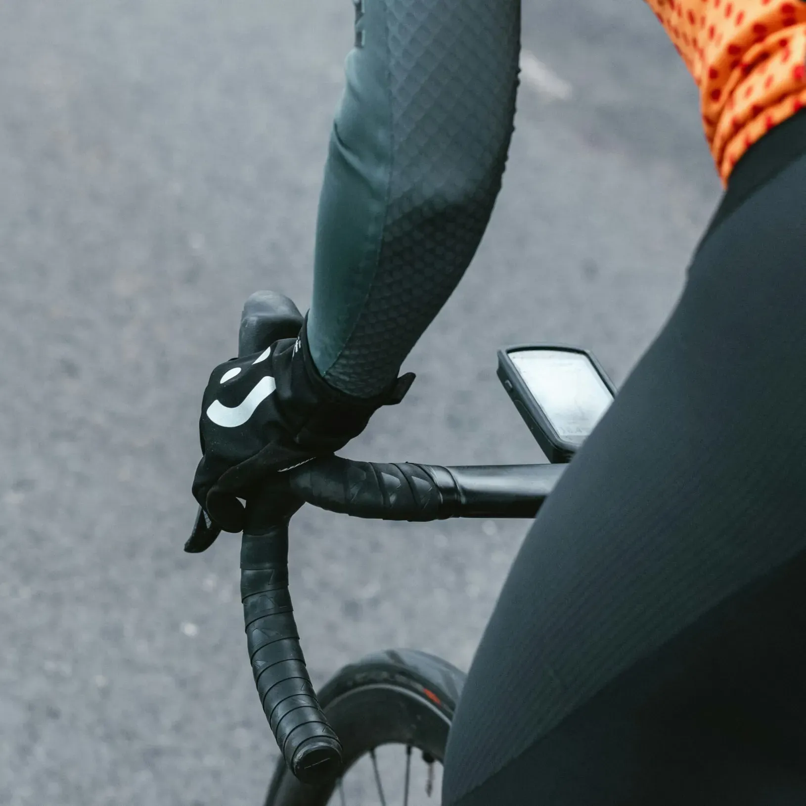Close-up of a road bike on the road with a cyclist pedaling standing up, wearing gloves, arm warmers, an orange t-shirt, and shorts; showing the road surface, front wheel, handlebar with onboard computer, and the cyclist’s hand and hip while pedaling.