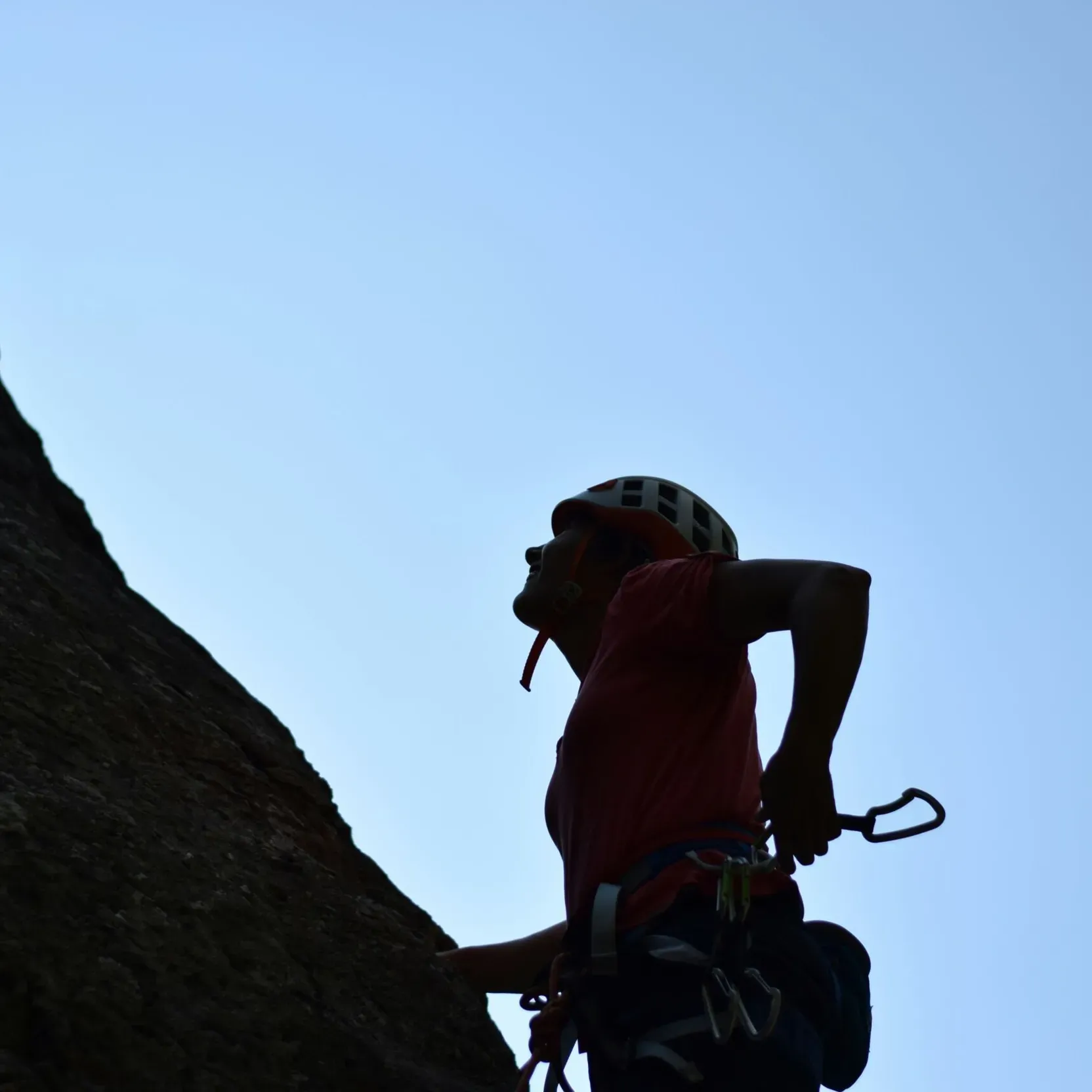 Silhouetted rock climber wearing a helmet, scaling a cliff under a clear blue sky with climbing gear, conveying determination and a sense of adventure.