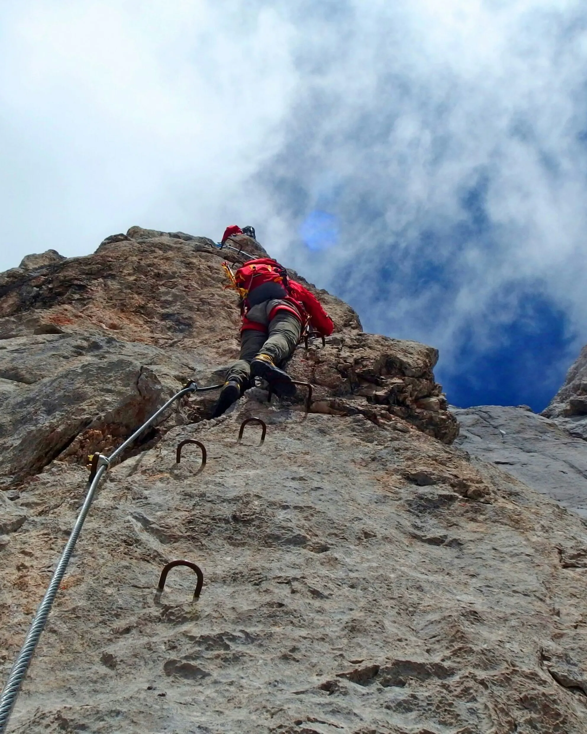 Climber in a red jacket ascending a steep rocky via ferrata with metal rungs, against a backdrop of blue sky and clouds, conveying a sense of challenge and adventure.