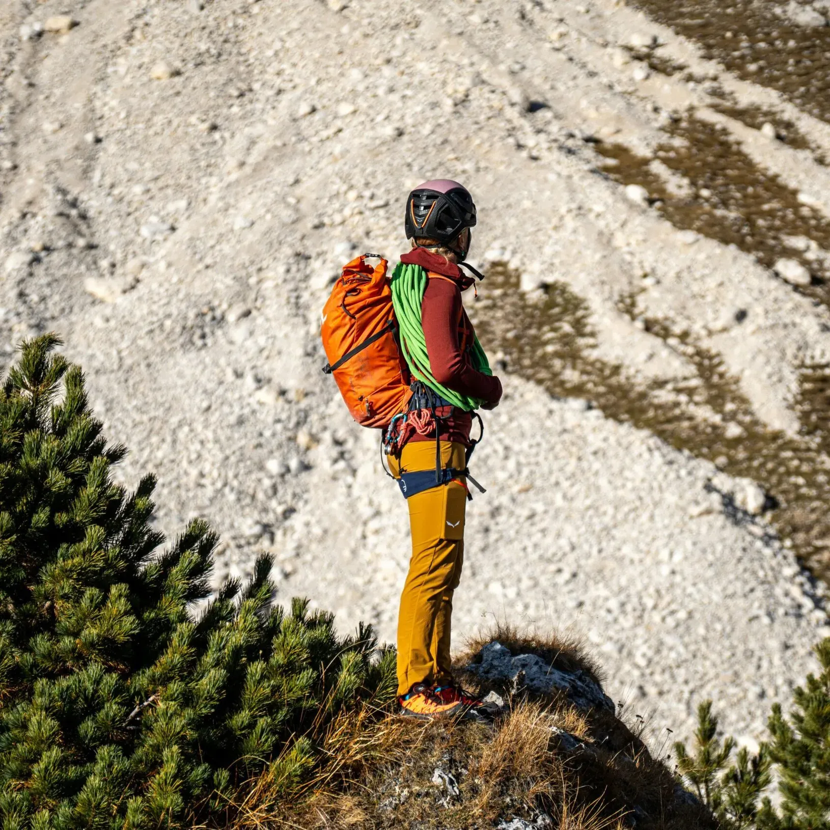 Hiker wearing a helmet, bright clothing, and an orange backpack standing on a grassy ledge, overlooking a sunlit rocky mountain landscape.